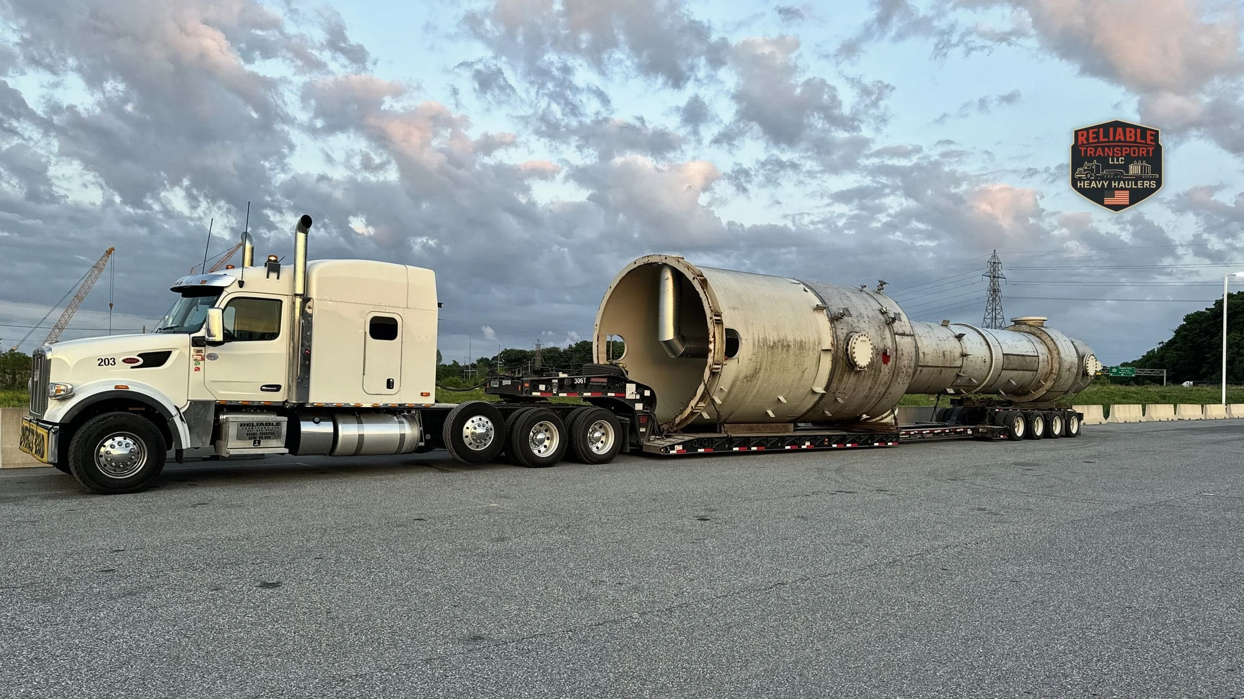 White heavy haul tractor pulling a large tan steel vessel on a multi axle trailer under an evening sky