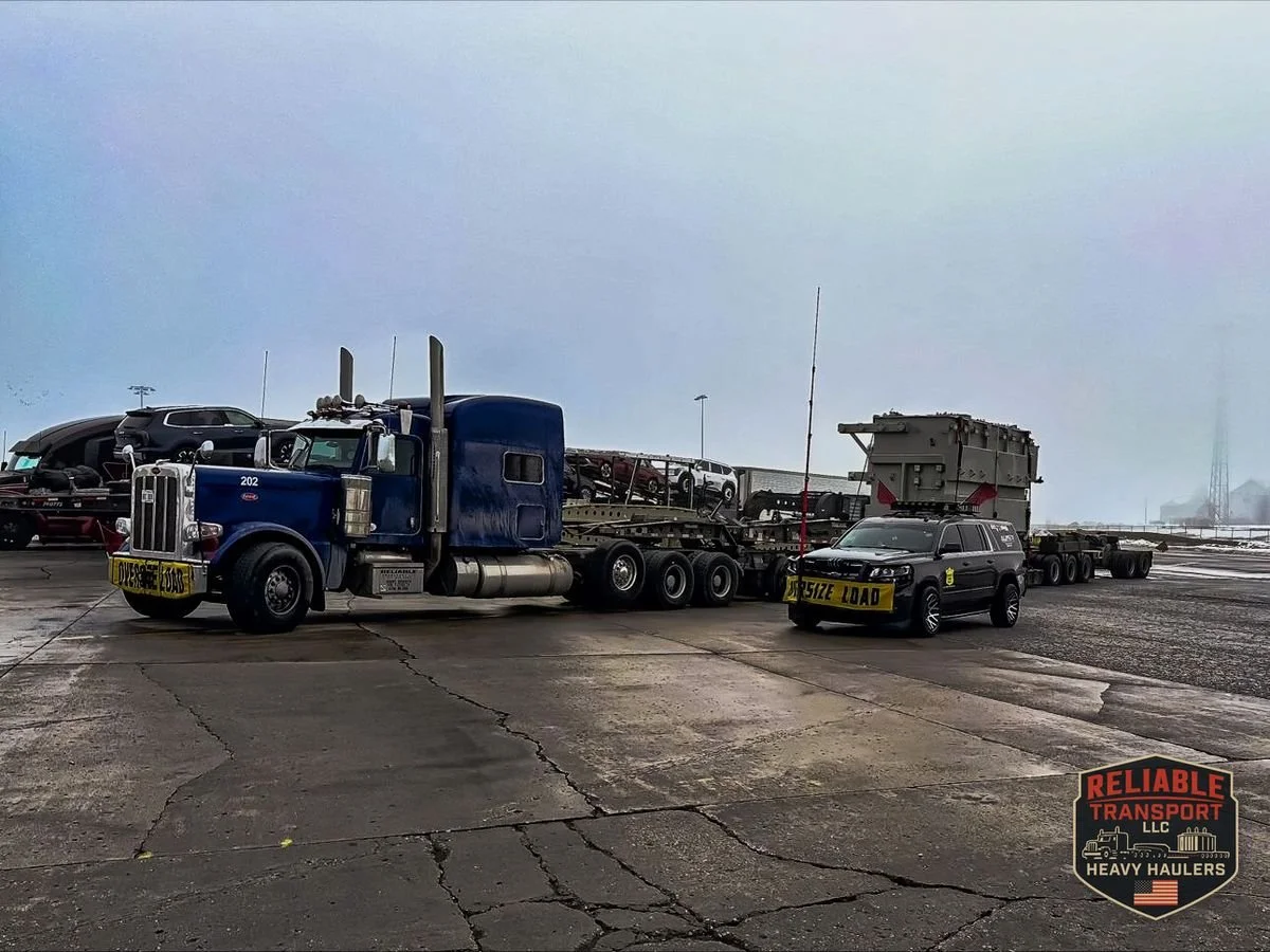 Blue heavy haul truck with a transformer on a multi axle trailer, parked beside an escort SUV with an OVERSIZE LOAD sign in a staging lot