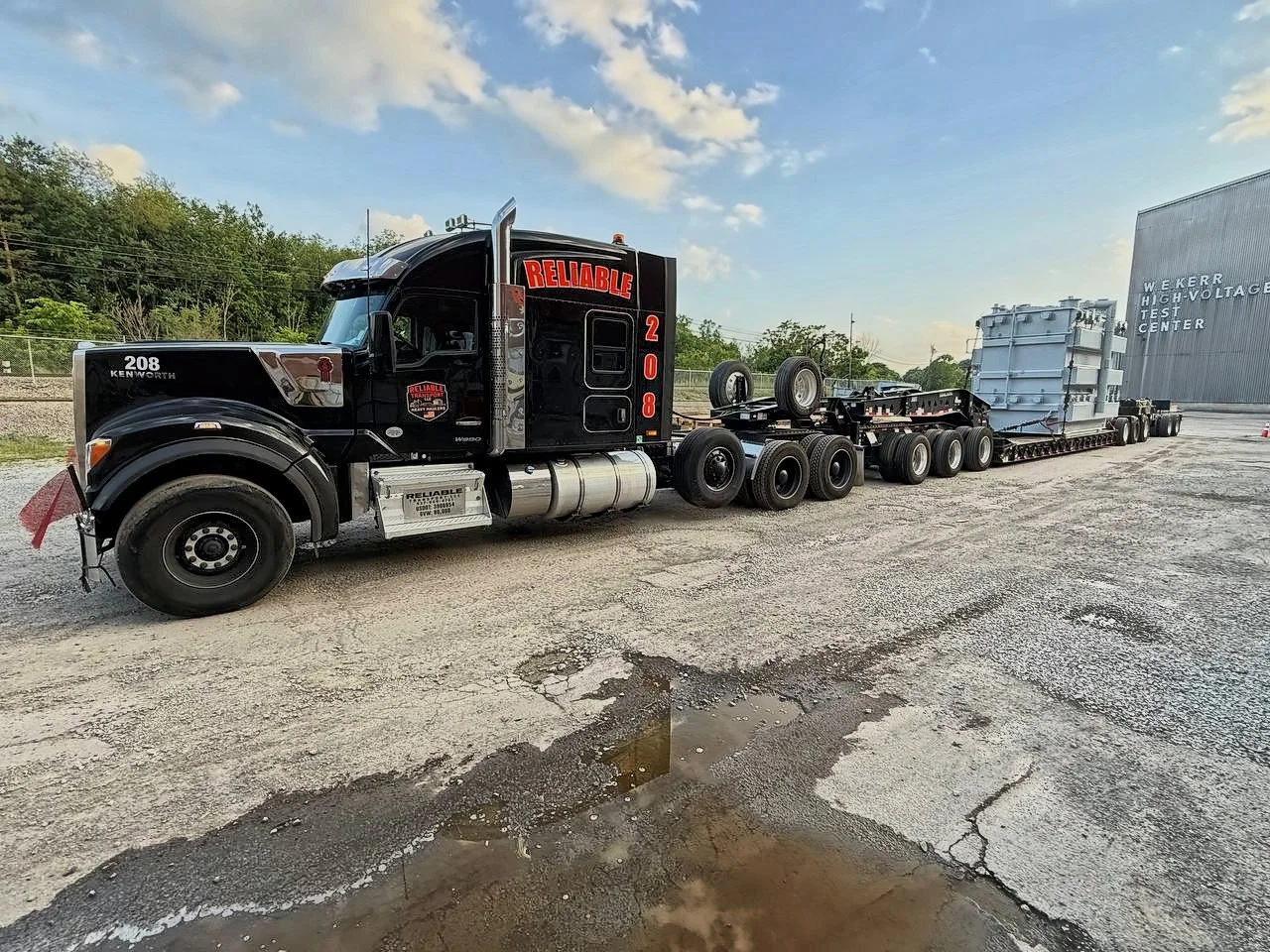Black Reliable truck 208 pulling a transformer on a multi axle trailer, trees and a building behind