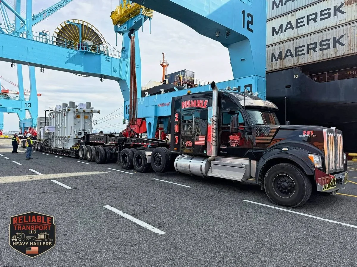Black Reliable truck 207 with a transformer on a multi axle trailer at a port, ship to shore cranes and stacked containers behind