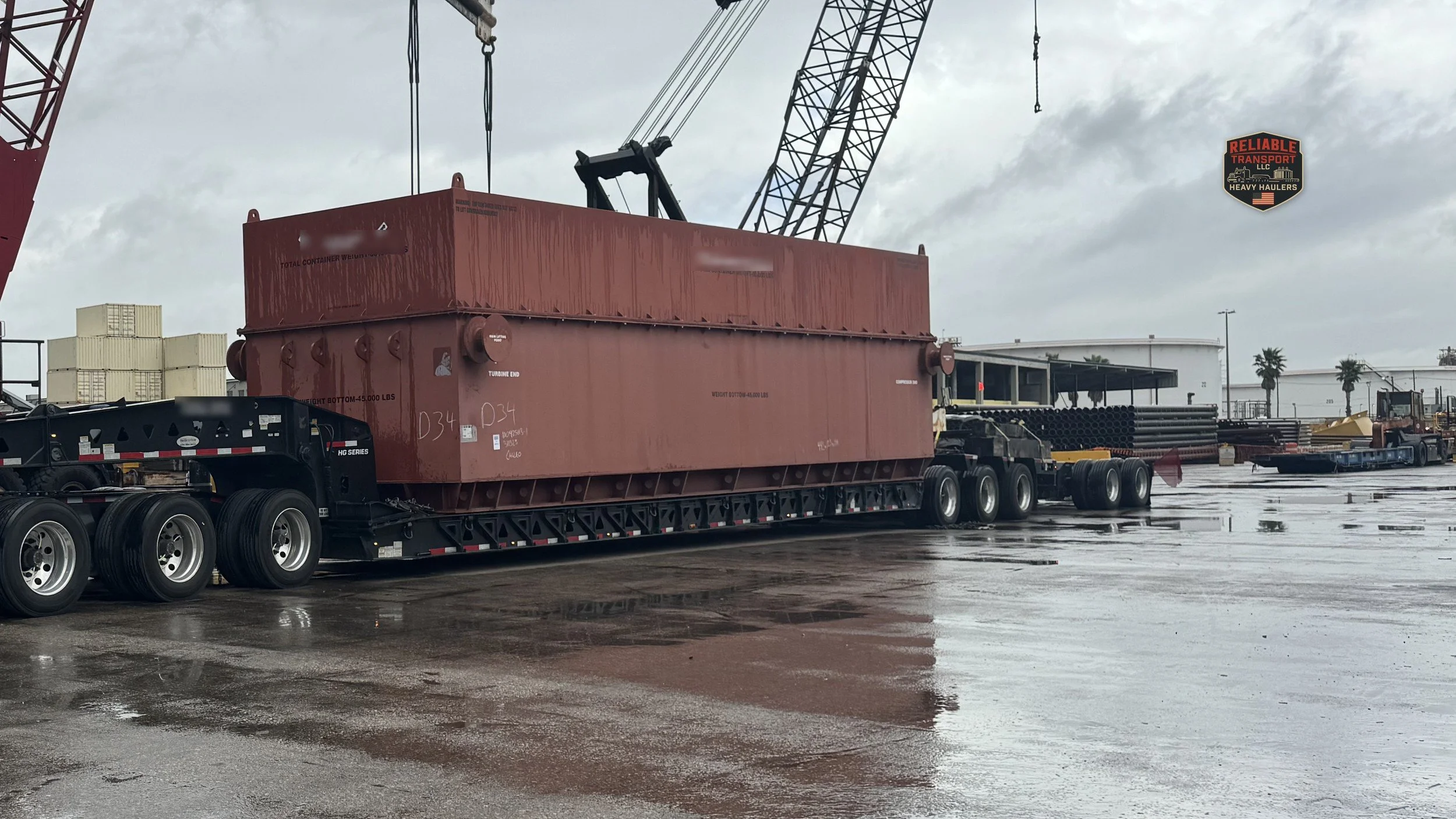 Large red steel box on a multi axle trailer below lattice boom cranes on a wet paved yard