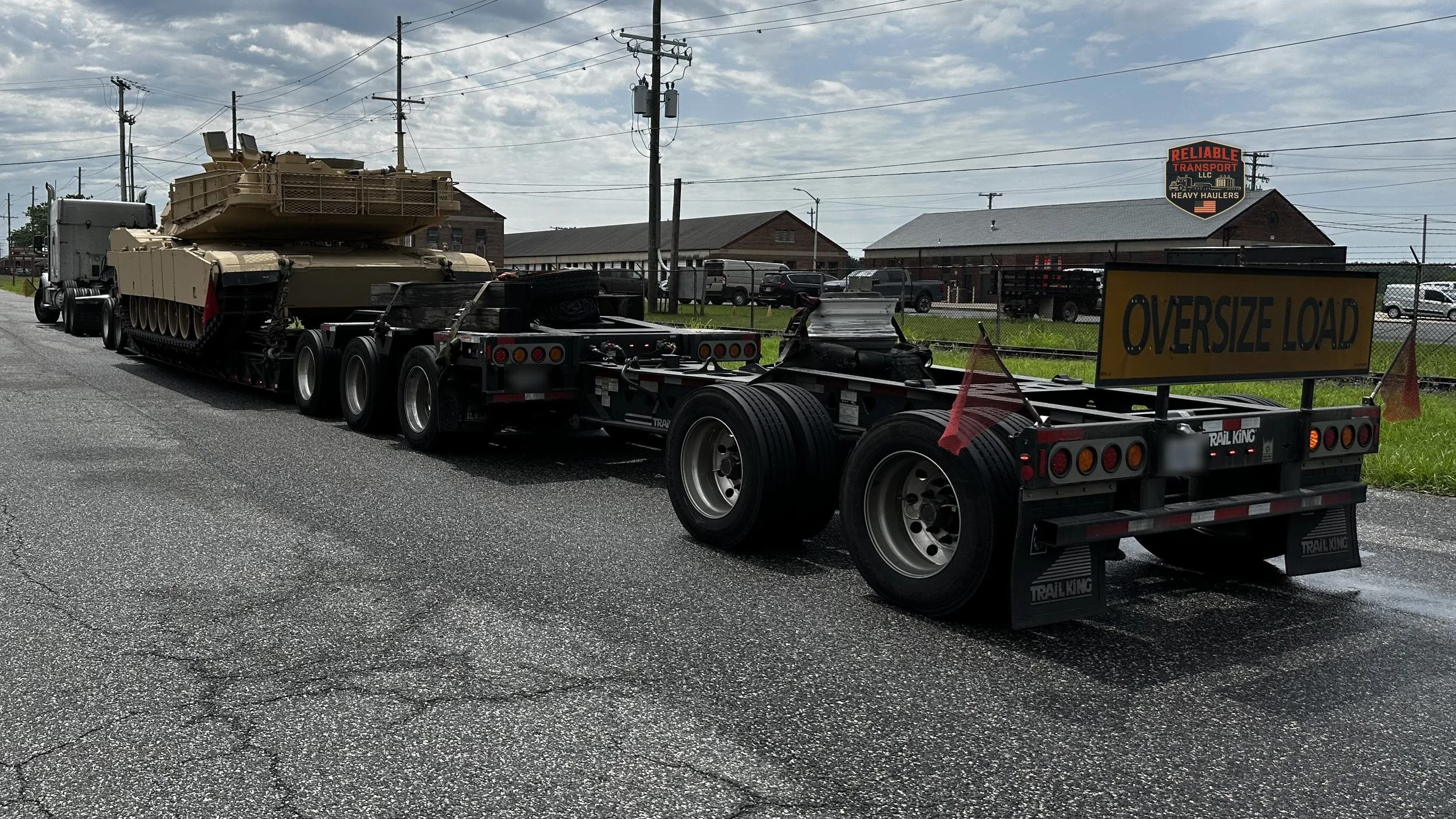 Tan tracked military vehicle on a multi axle trailer with an OVERSIZE LOAD banner, seen from behind on a two lane road under a cloudy sky