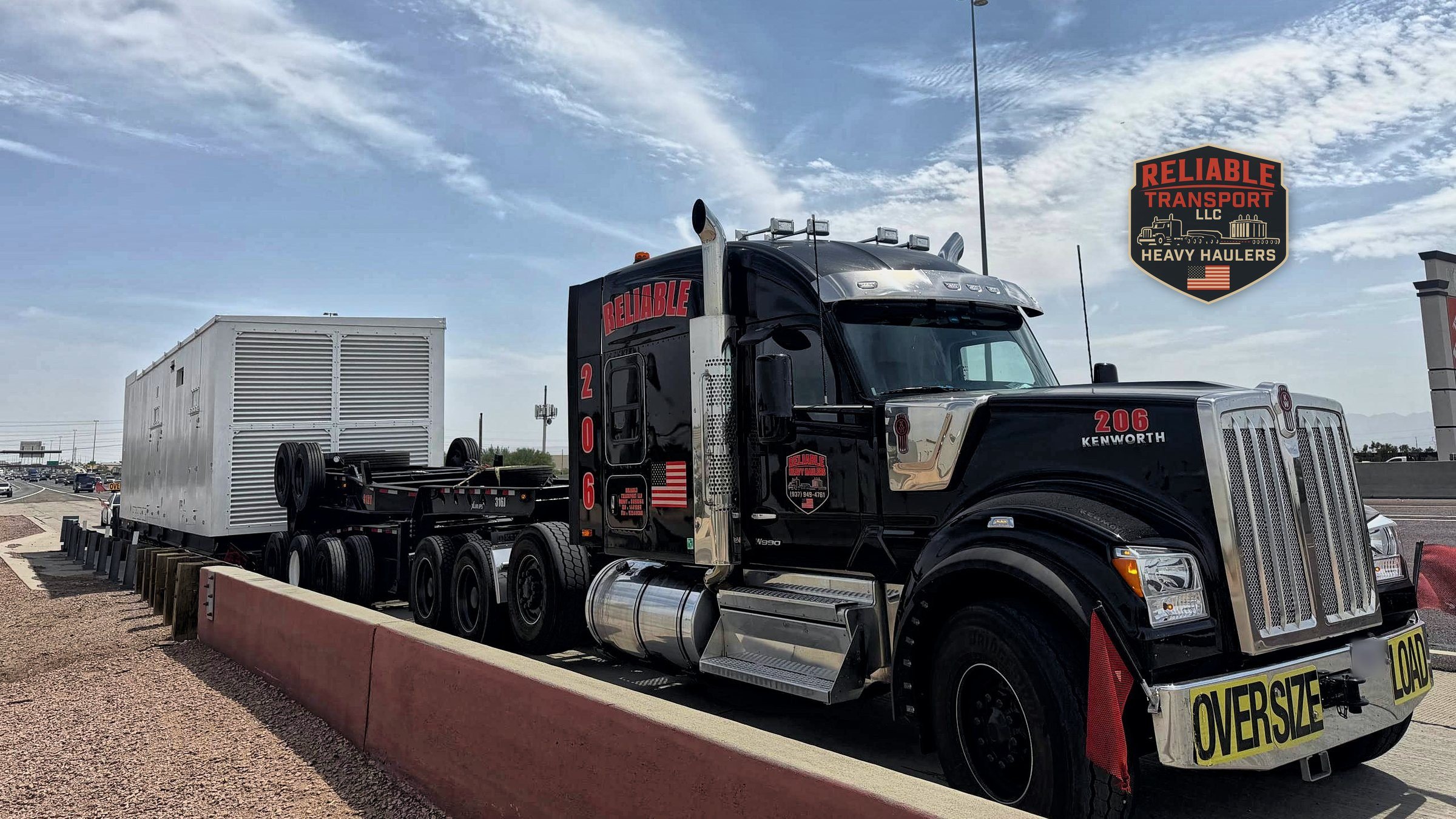 Black heavy haul truck, unit 206, with an OVERSIZE LOAD banner and a large white enclosure on a multi axle trailer under a big sky