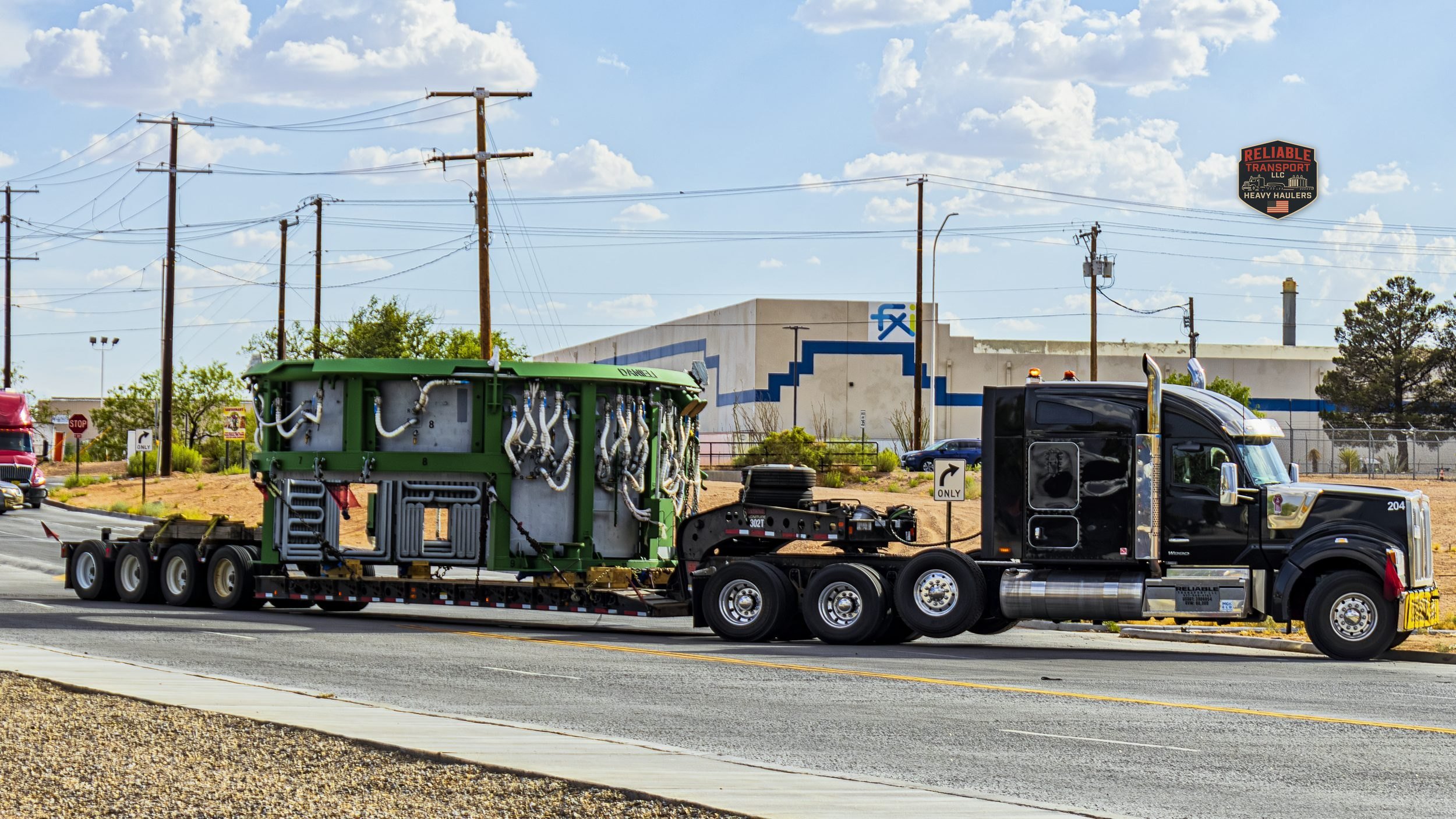 Black heavy haul tractor pulling a green industrial unit on a multi axle trailer along a road beside a warehouse