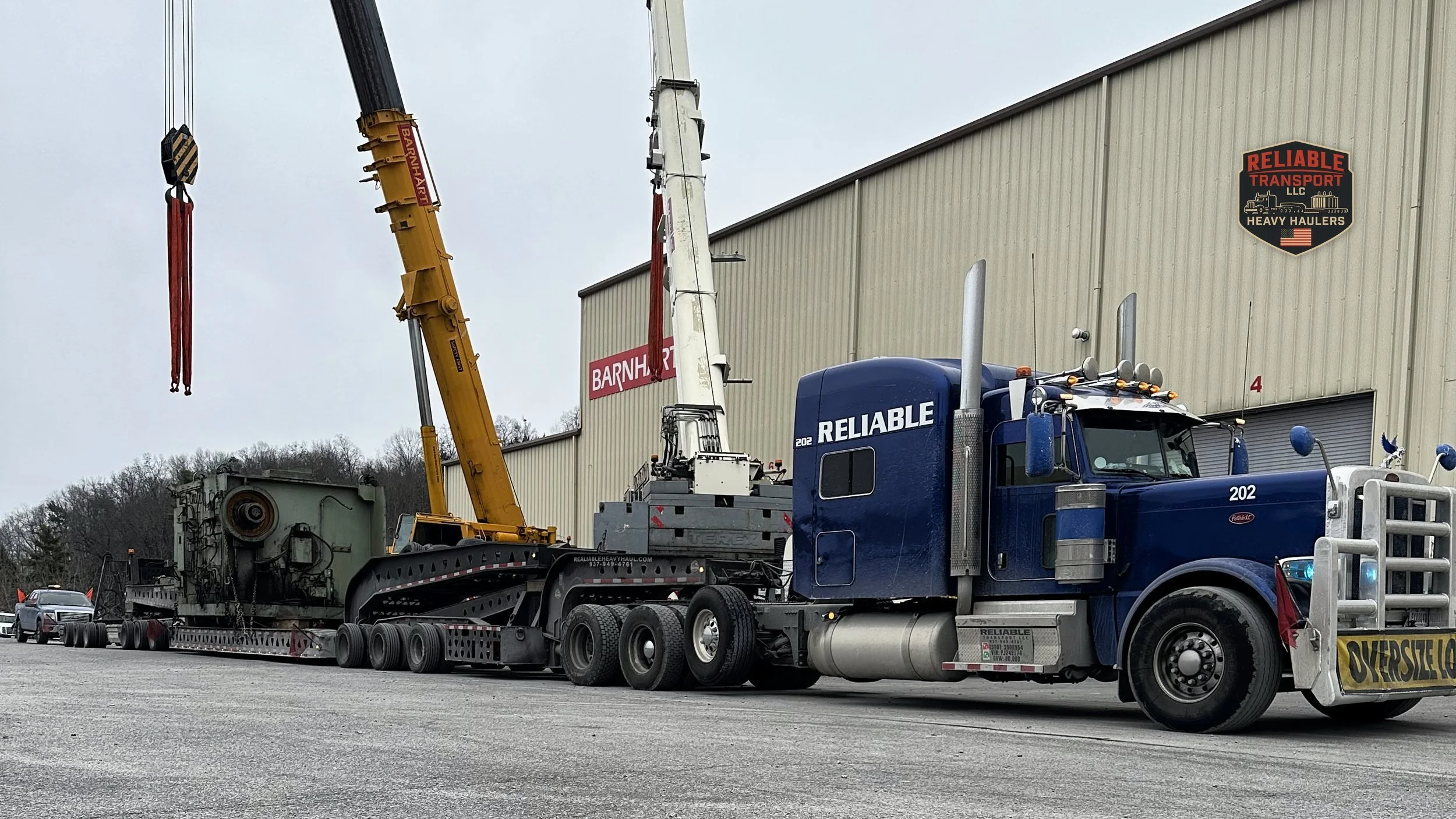 Blue heavy haul tractor with a large industrial load on a multi axle trailer beside two cranes and a warehouse
