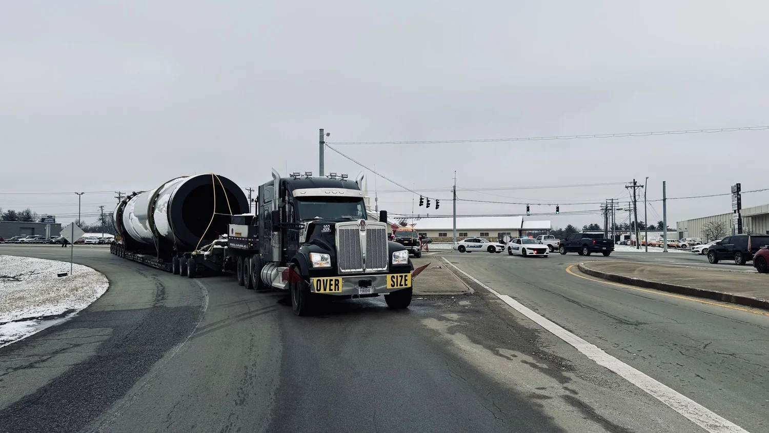 Black heavy haul truck with an OVERSIZE banner pulling the dryer drum through an intersection while police cars hold traffic