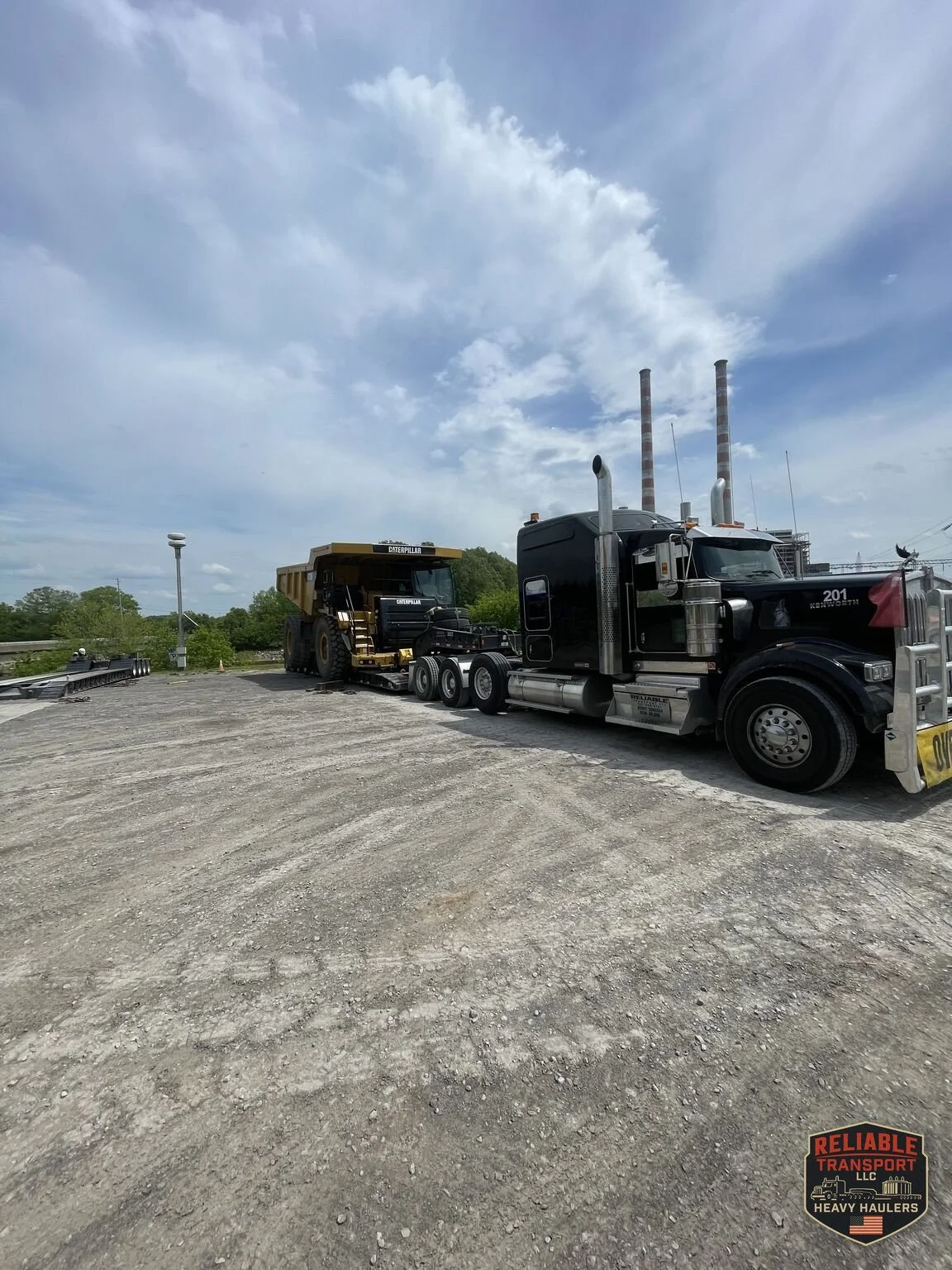 Black heavy haul truck with a loaded multi axle trailer parked in a wide gravel lot under a tall white cloud