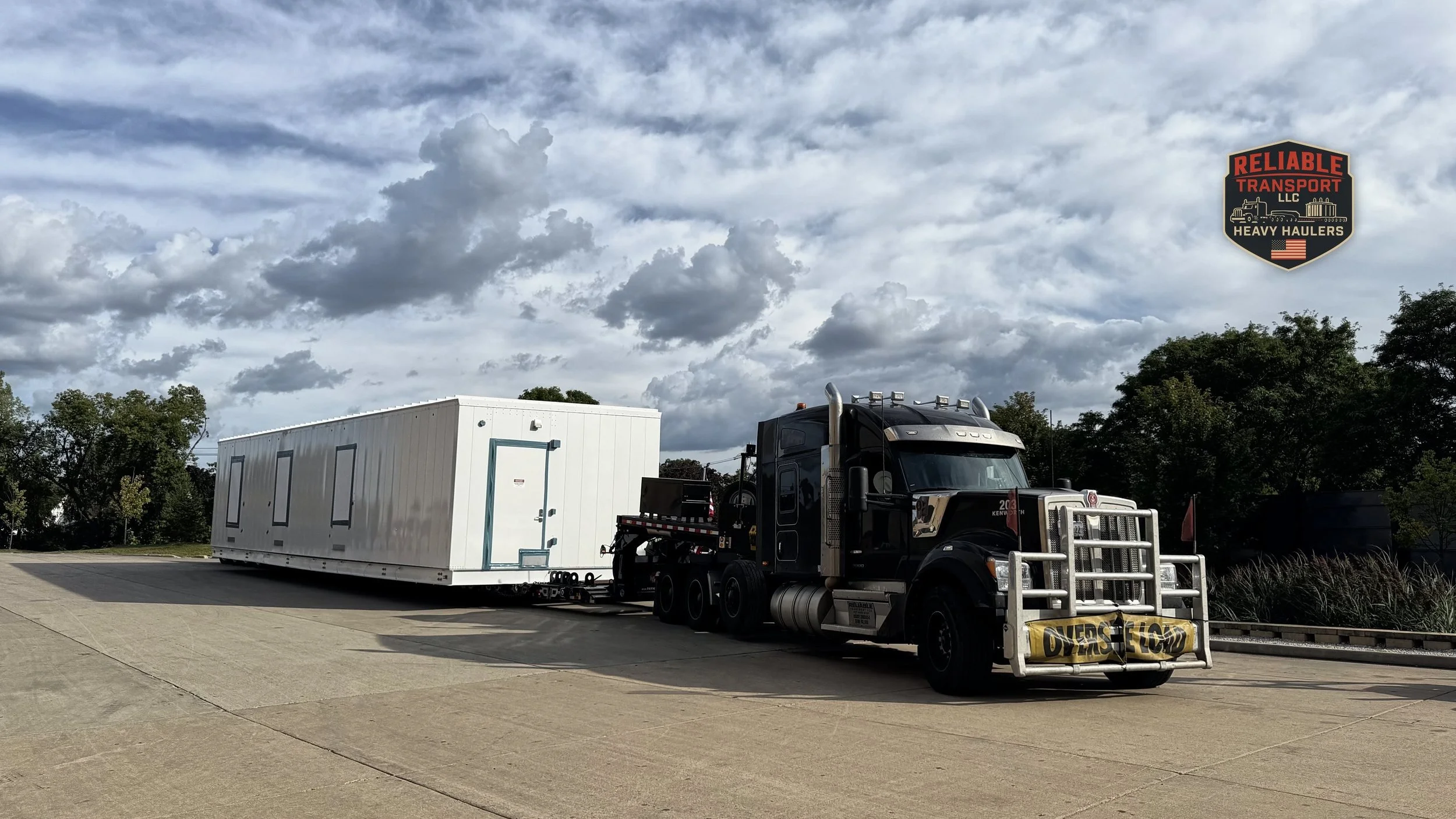 Black heavy haul tractor pulling a white modular building on a trailer under a cloudy sky