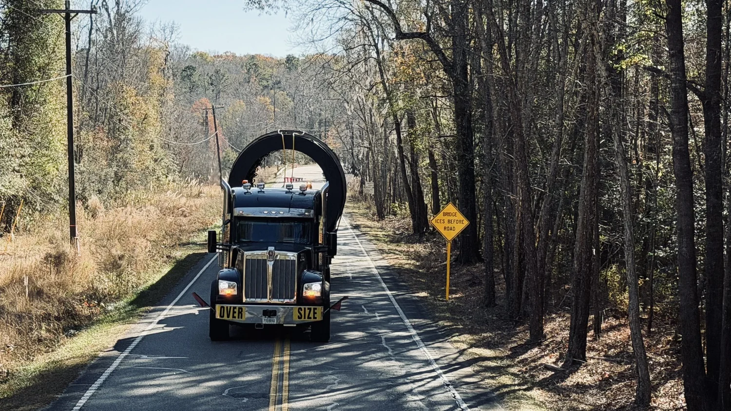 Front view of the truck hauling the dryer drum down a two lane road through the woods, past a Bridge Ices Before Road sign