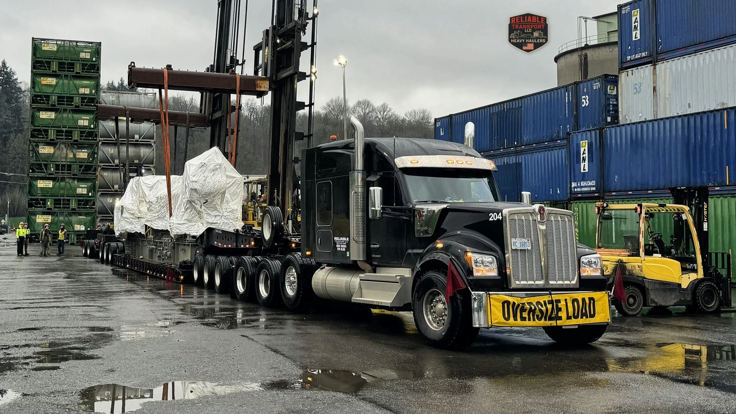 Black heavy haul tractor pulling a tarped load on a multi axle trailer in a wet container yard beside a forklift