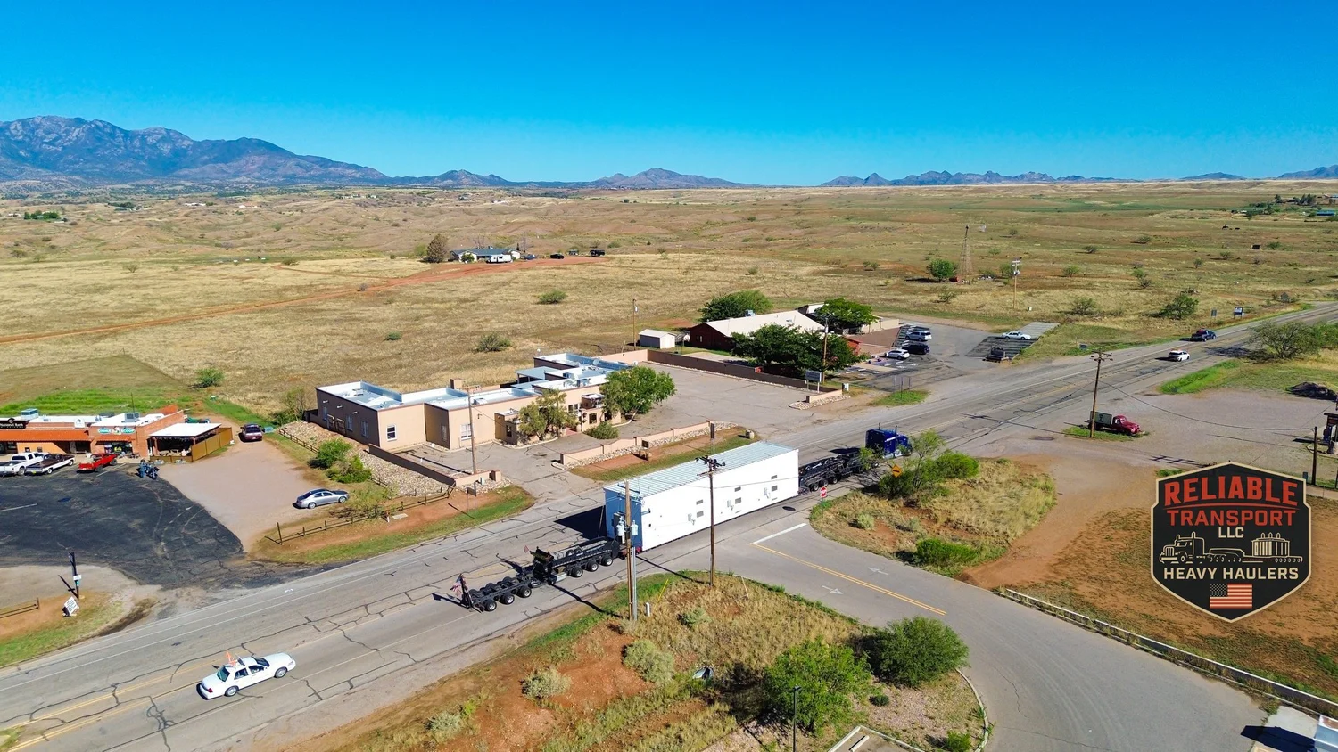 Drone view of the white switchgear building and a blue truck turning through a small town intersection, with an escort car on the road