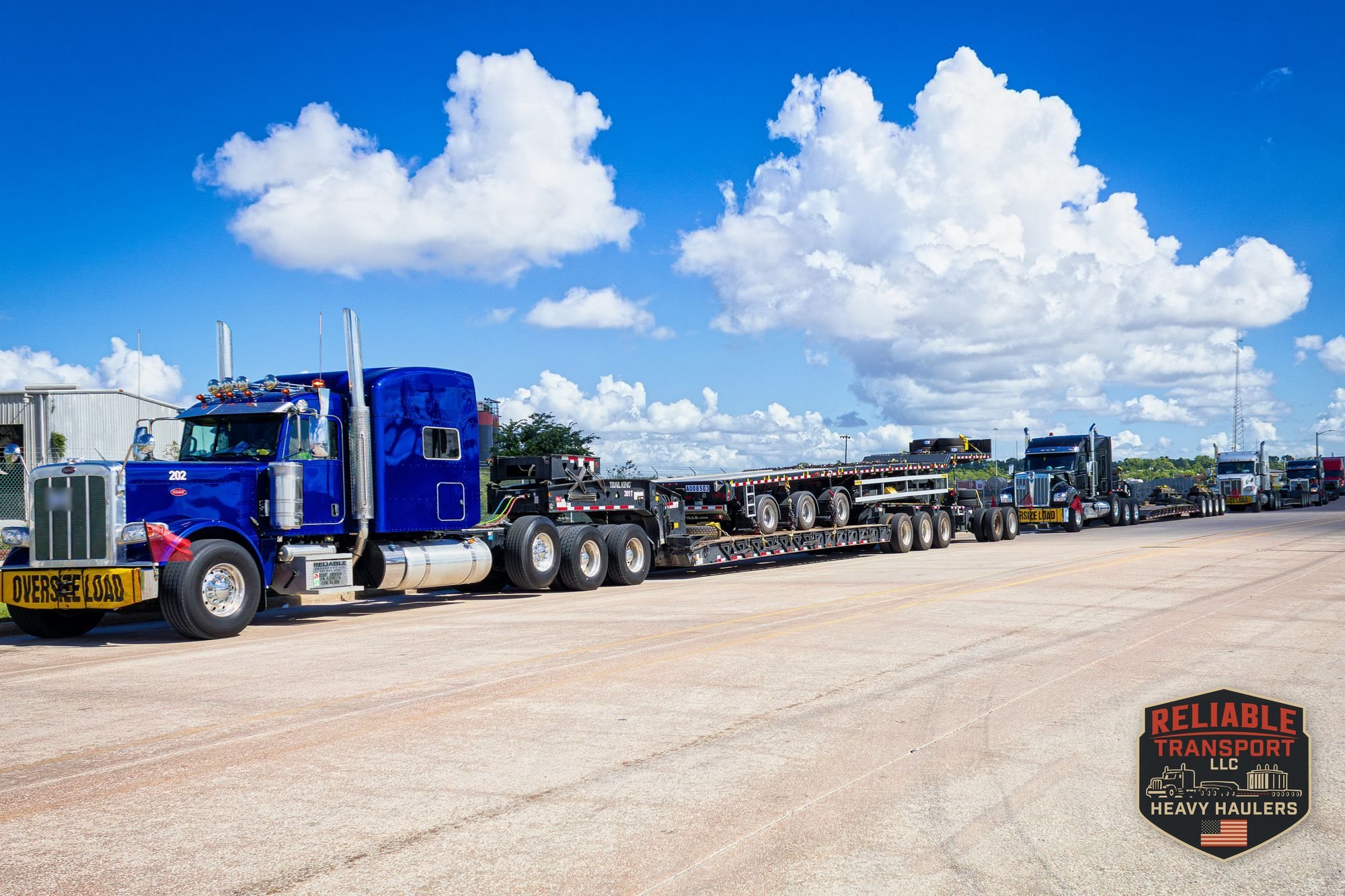 Blue heavy haul truck leading a line of trucks with multi axle trailers staged in a lot under big white clouds