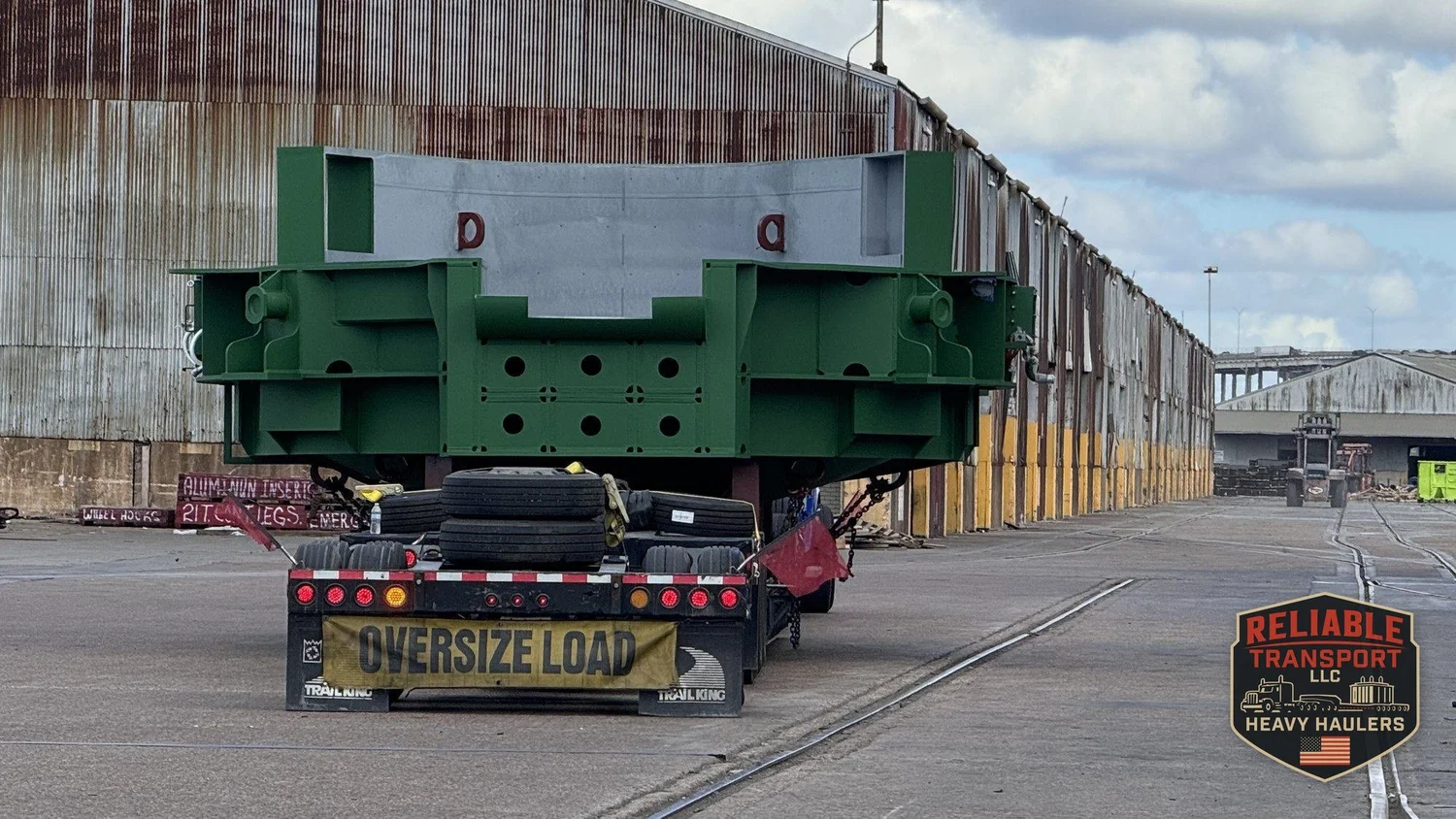 Wide green load on a multi axle trailer with an OVERSIZE LOAD banner in a port yard beside a long warehouse