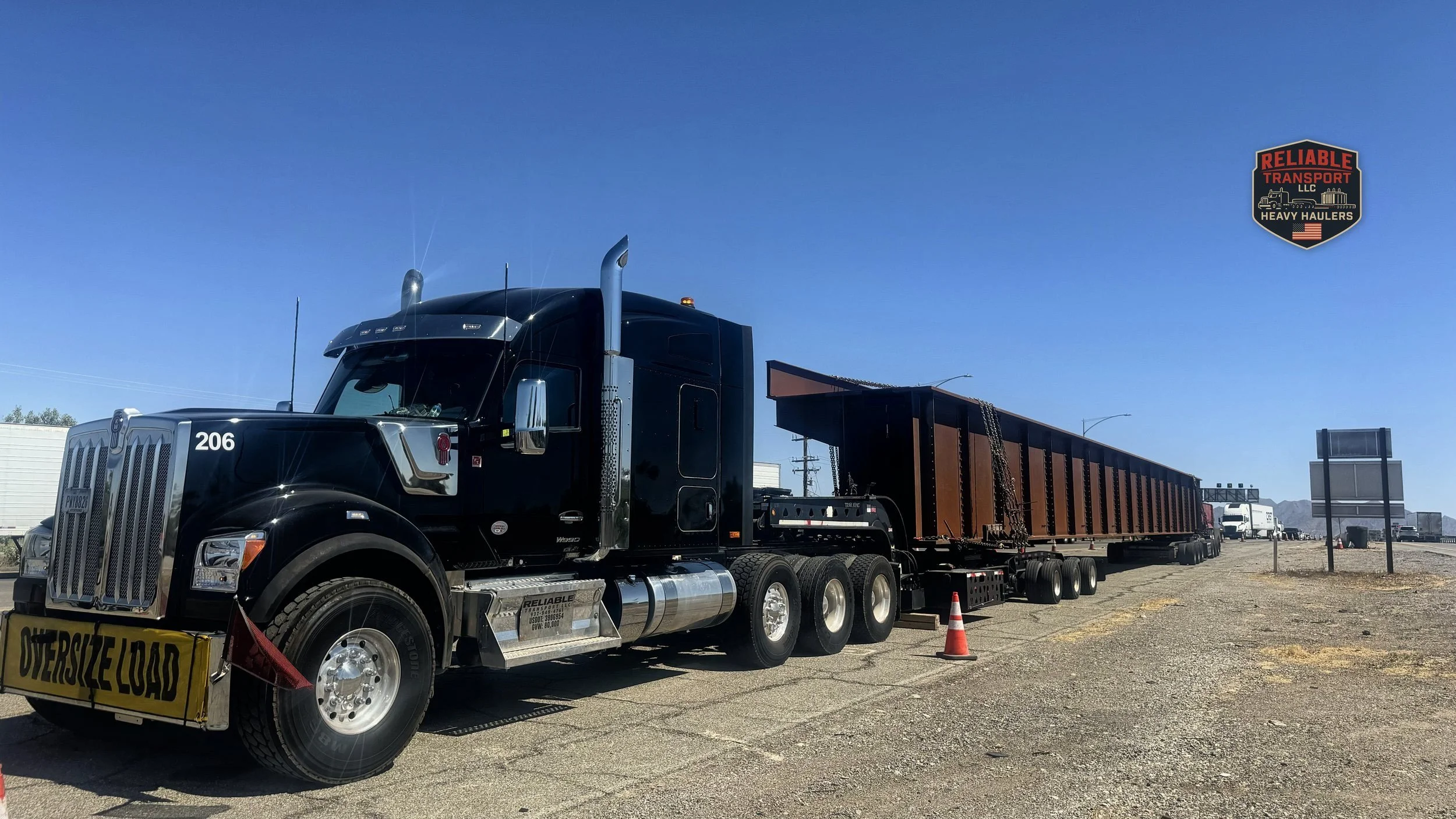 Black heavy haul tractor with an OVERSIZE LOAD banner pulling a long rust colored steel load on a multi axle trailer under a clear sky