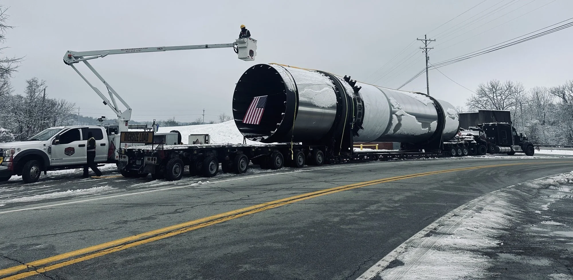Bucket truck crew lifting overhead lines as a long dryer drum with an American flag on its open end rolls along a snowy road on a heavy haul trailer