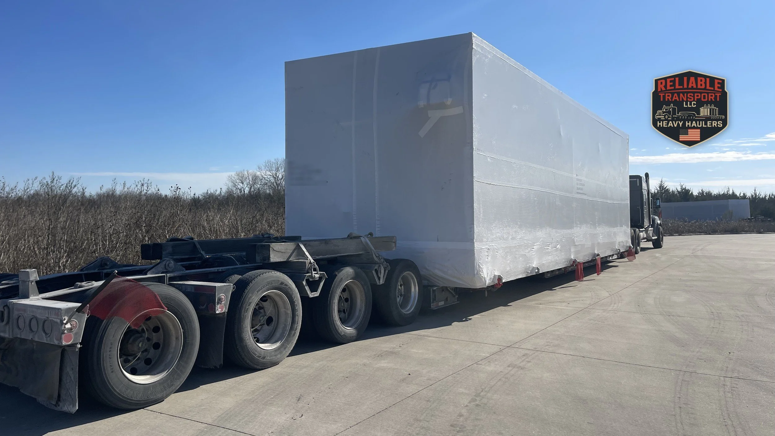 Large white wrapped module on a multi axle trailer in a paved yard under a clear blue sky
