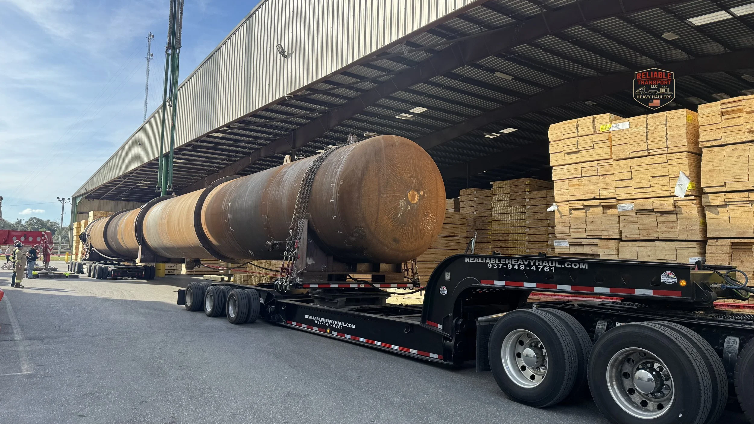 Long rusted steel vessel on a multi axle trailer beside stacked lumber under a warehouse canopy