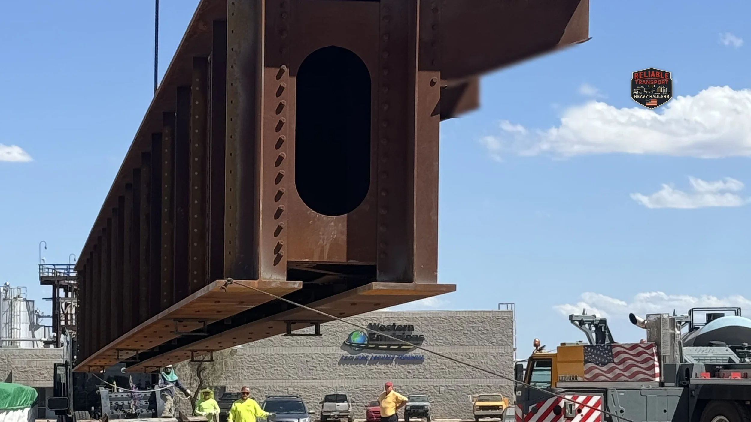 Steel module suspended from a mobile crane over a multi axle trailer in a paved yard under a clear sky