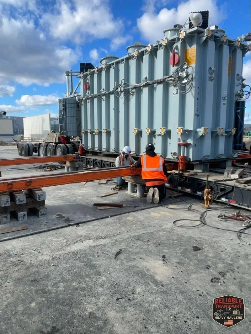 The transformer set on orange skid beams with a crew member in an orange vest working beside it under a blue sky