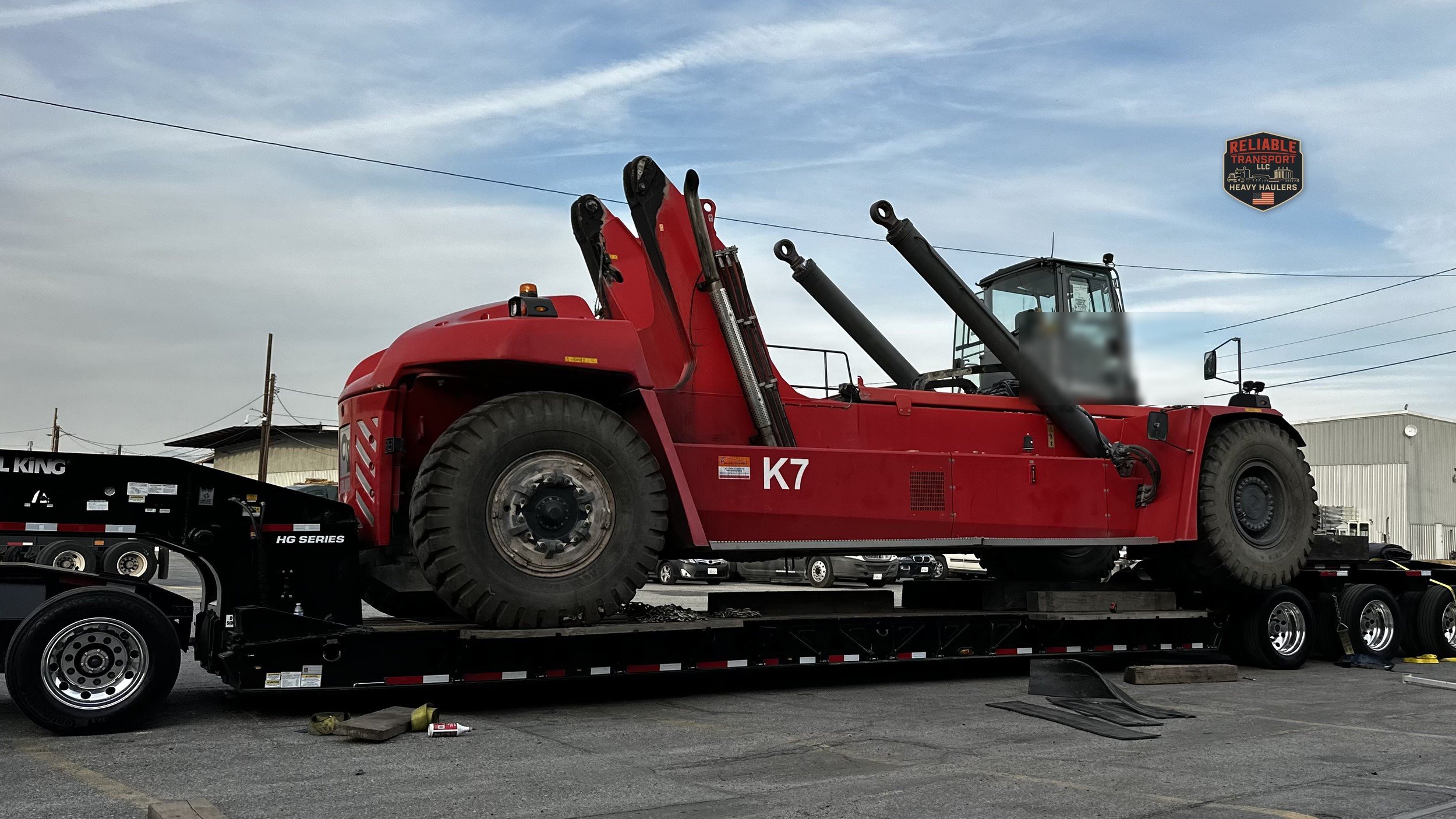 Red container handler lifting a steel load beside a multi axle trailer in an industrial yard