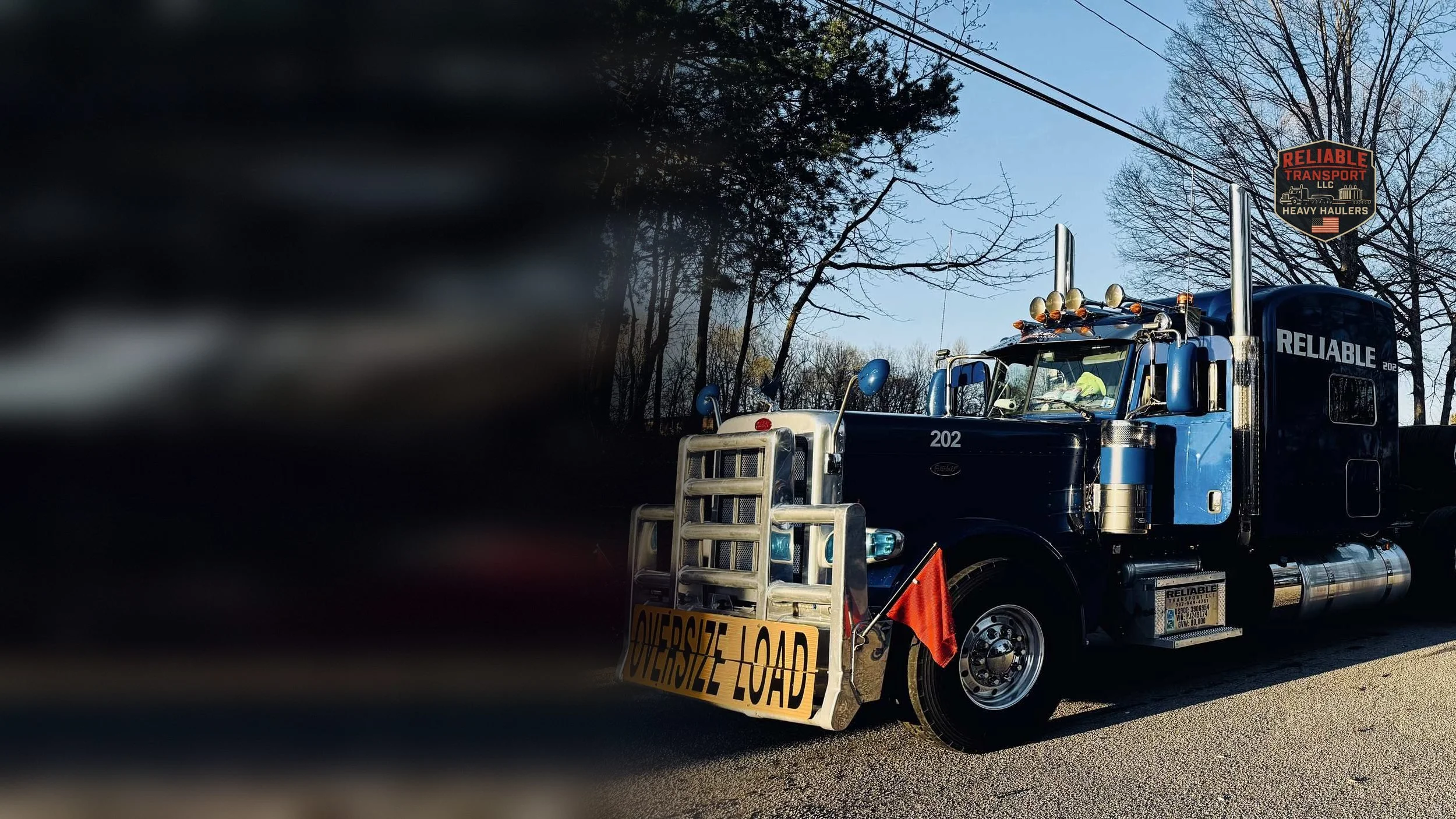 Blue heavy haul tractor with an oversize load banner on a tree lined road in late day sun