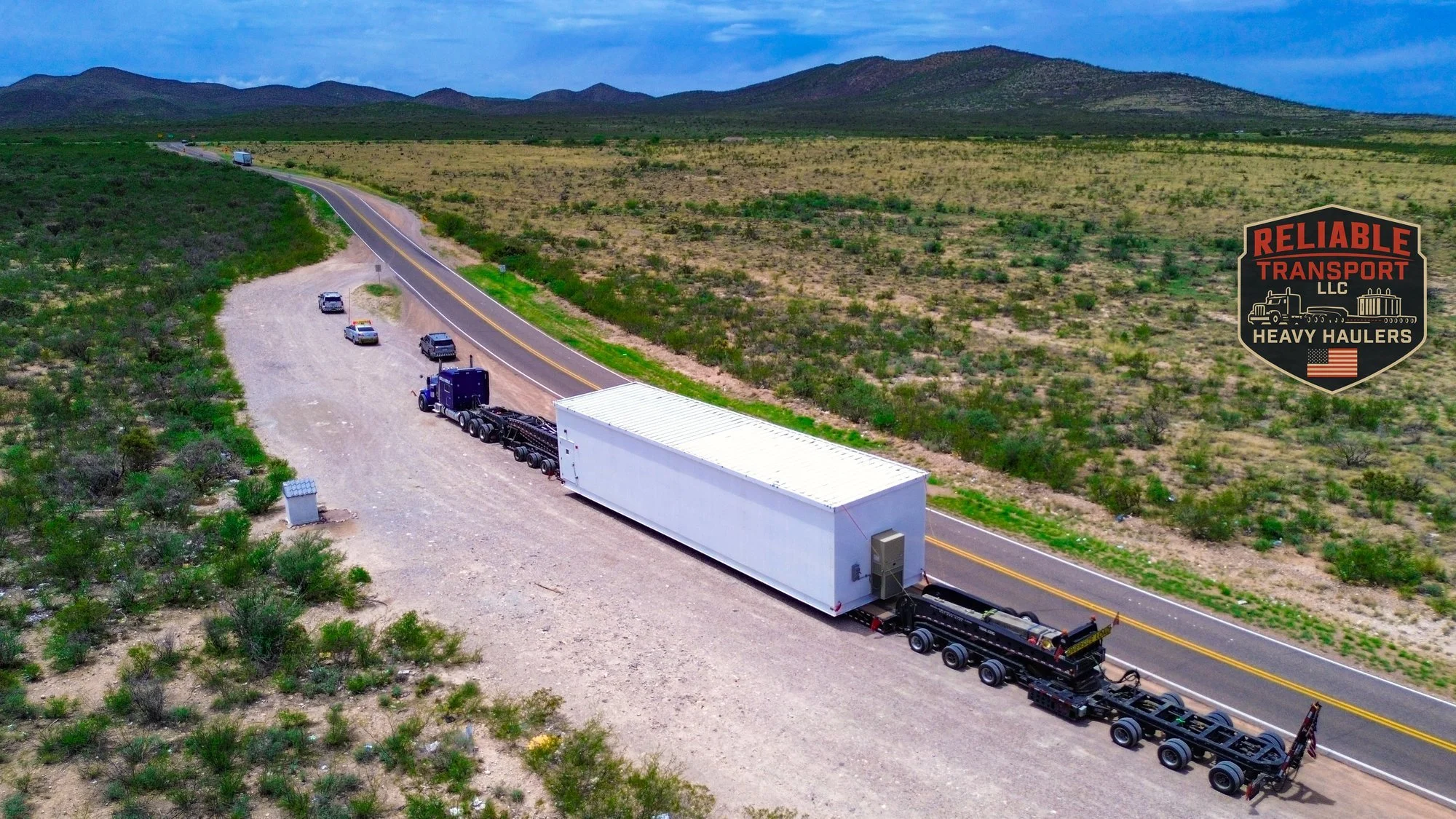 Drone view of a white switchgear building on a heavy haul trailer with steerable rear dollies, escort vehicles lined up ahead on a desert highway