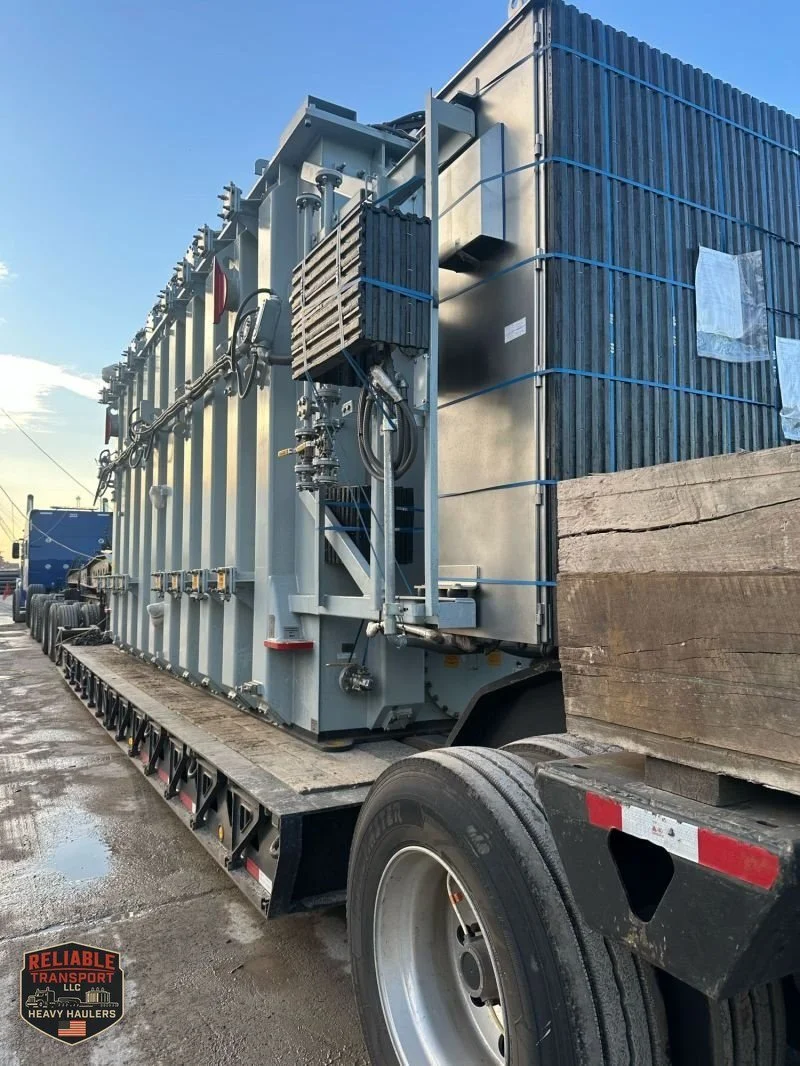 Side view of the gray power transformer loaded on the heavy haul trailer, with the truck in the foreground
