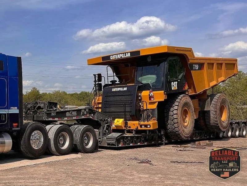 Yellow CAT 775F haul truck loaded on a multi axle trailer in a gravel yard under a blue sky