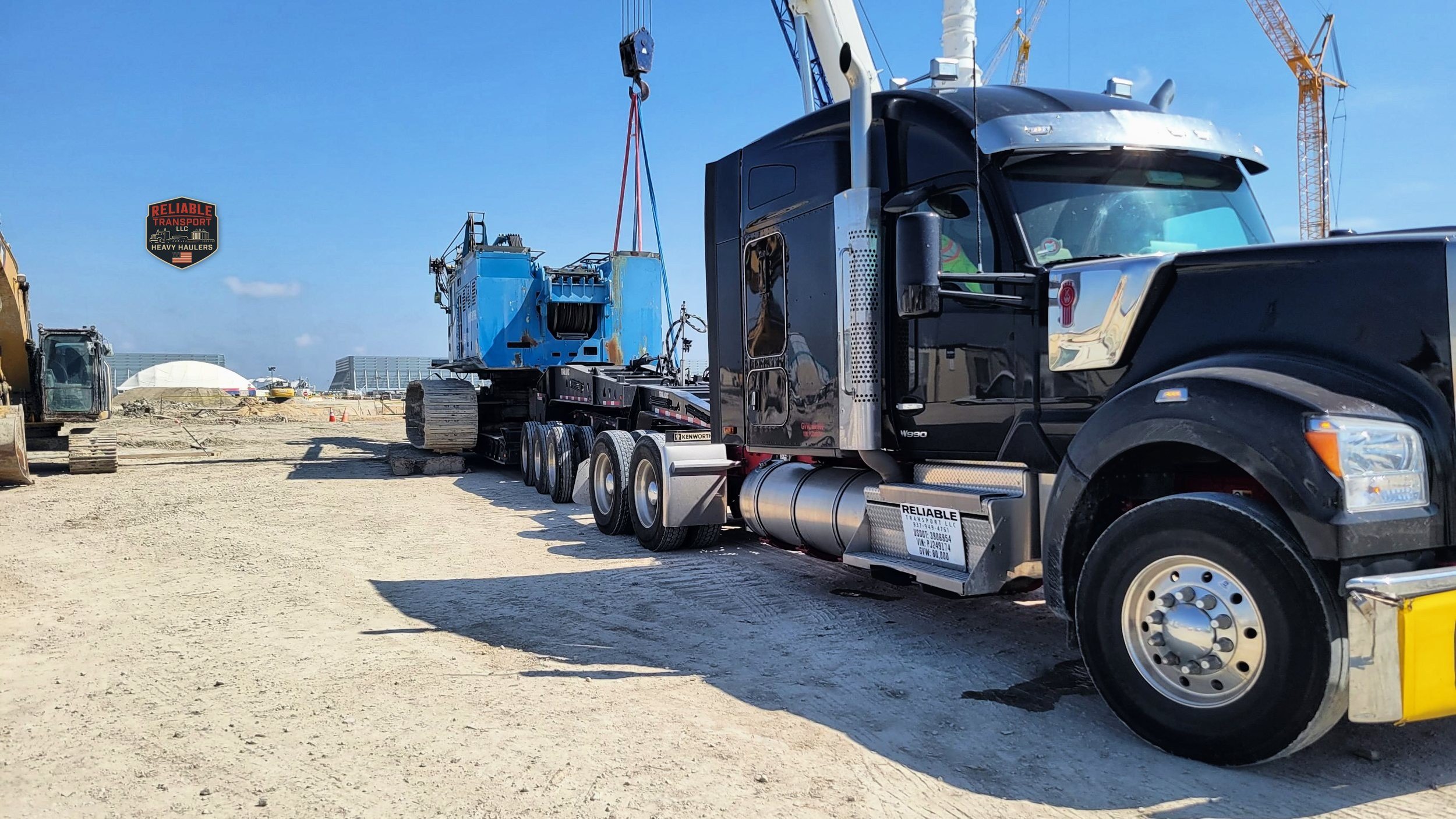 Black heavy haul tractor beside a blue crawler crane body on its multi axle trailer at a work site while a crane hook hangs above