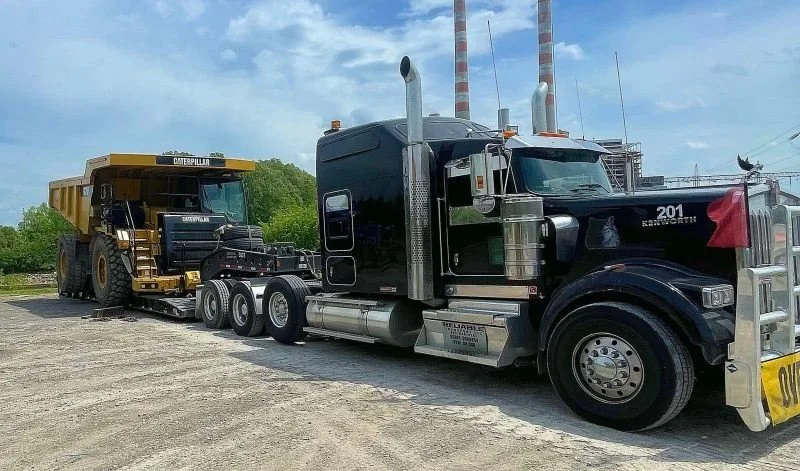Black heavy haul truck pulling a yellow CAT 775F haul truck on a multi axle trailer across a gravel lot