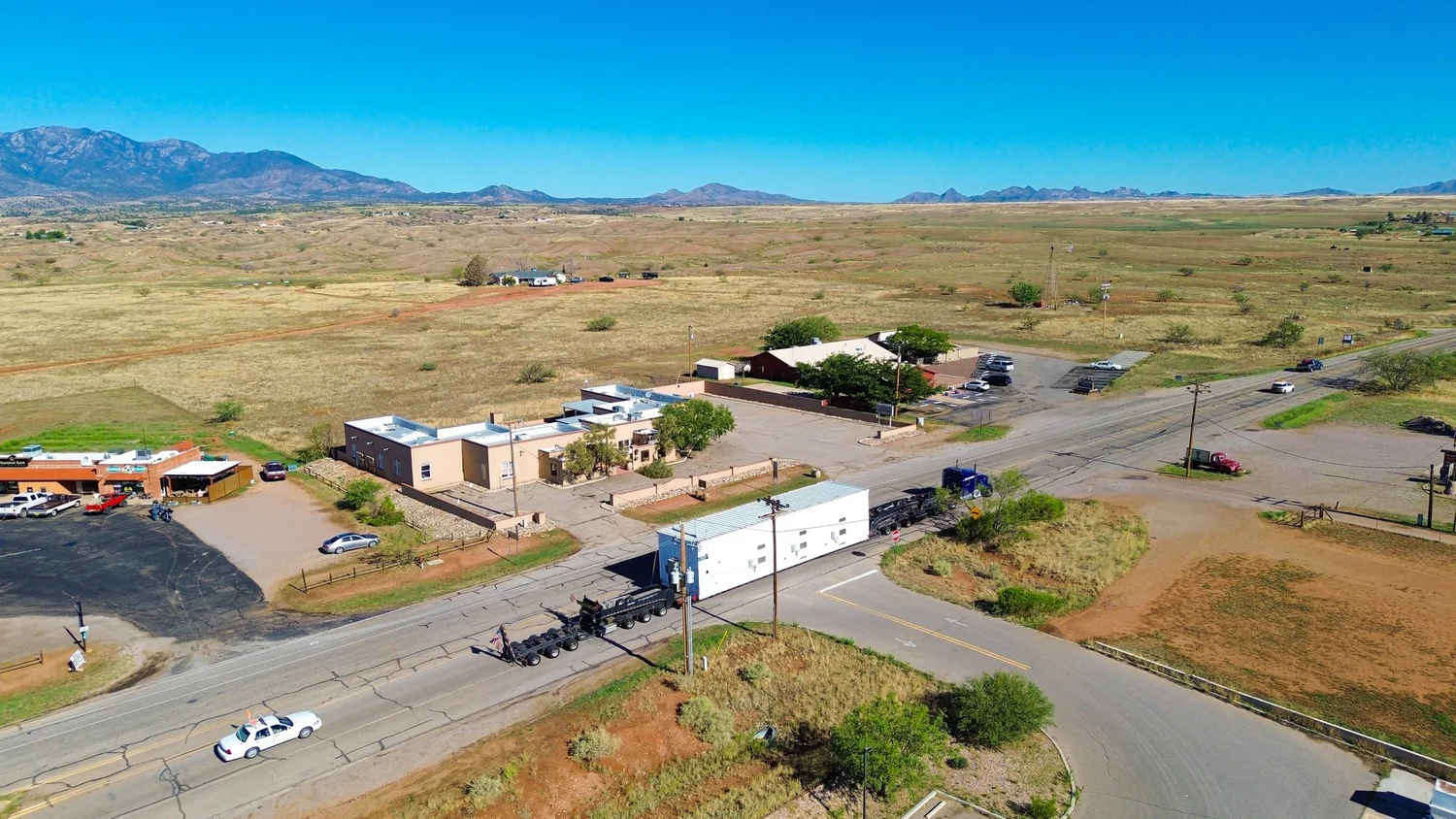 Drone view of the switchgear building on its trailer turning through a small town intersection, with an escort car on the road ahead