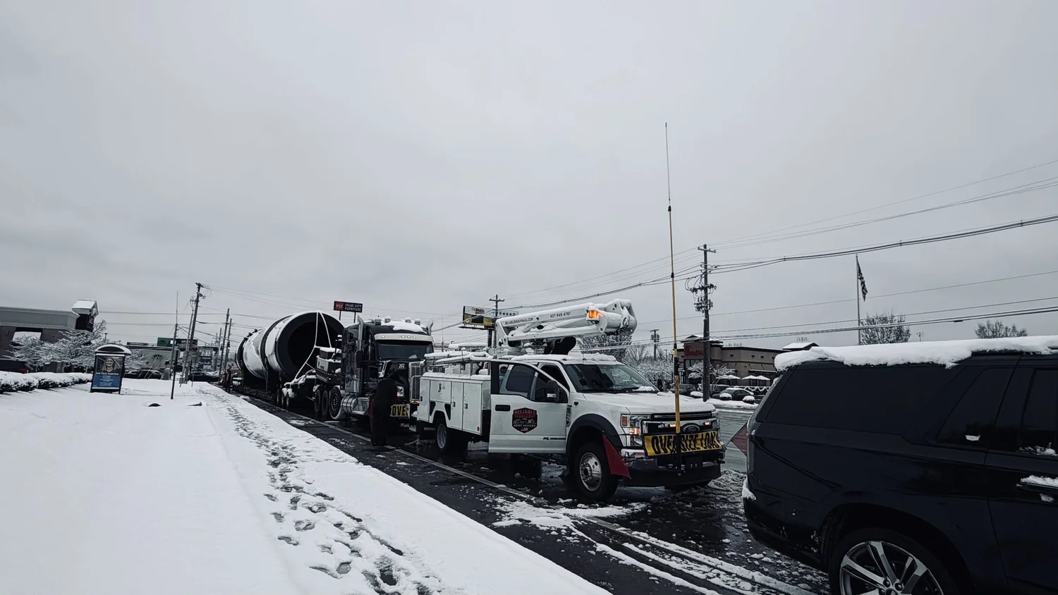 Dryer drum and truck stopped on a snowy street beside a white bucket truck carrying a tall high pole, with an escort vehicle alongside