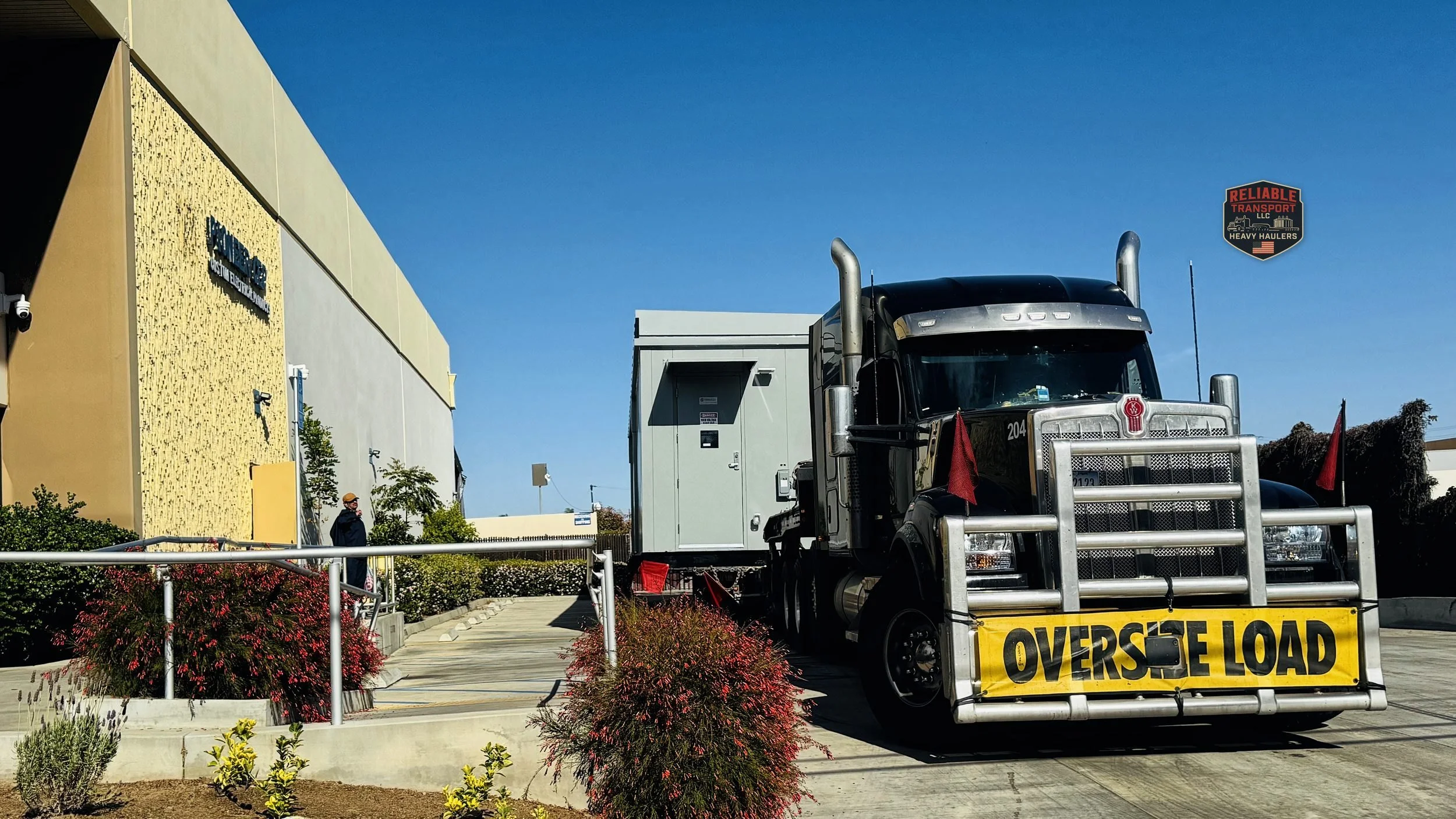 Black heavy haul tractor with an OVERSIZE LOAD banner pulling a gray modular building past an office building under a blue sky