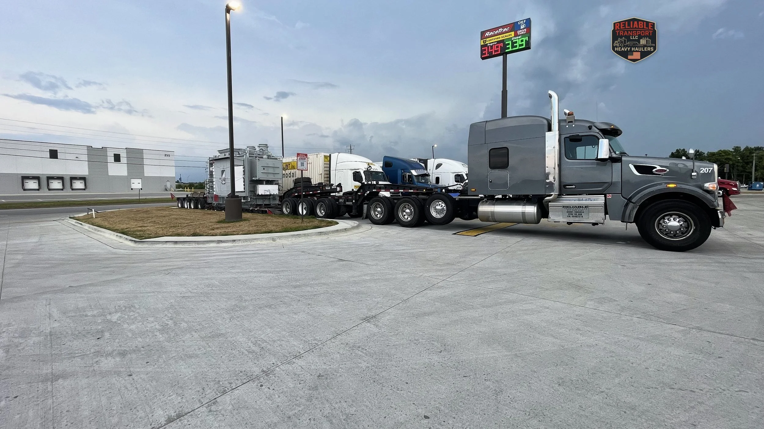 Gray heavy haul tractor pulling a tall gray industrial load on a multi axle trailer across a concrete lot under a cloudy sky
