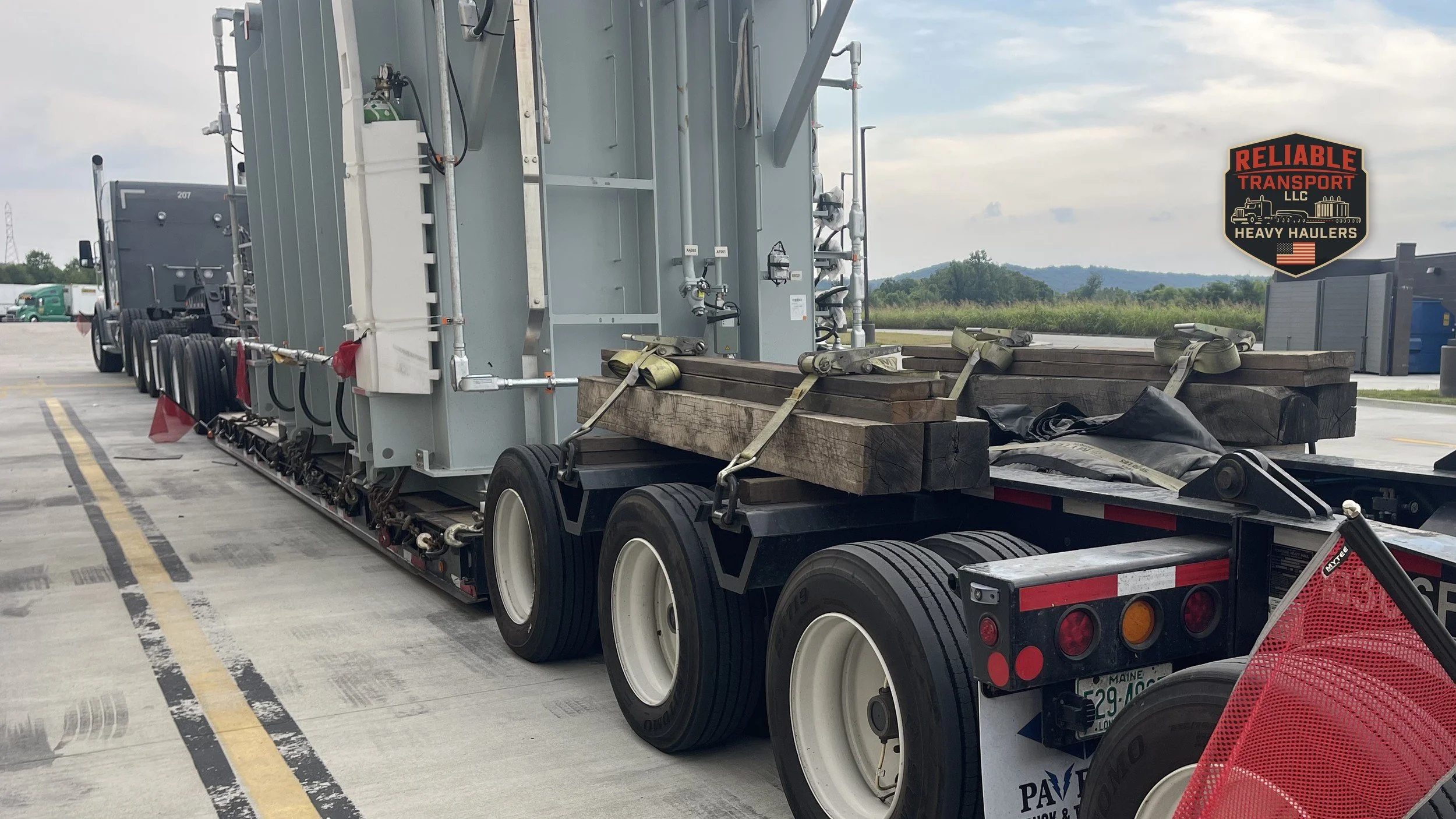 Large gray transformer secured on a multi axle trailer in a gravel yard with workers nearby