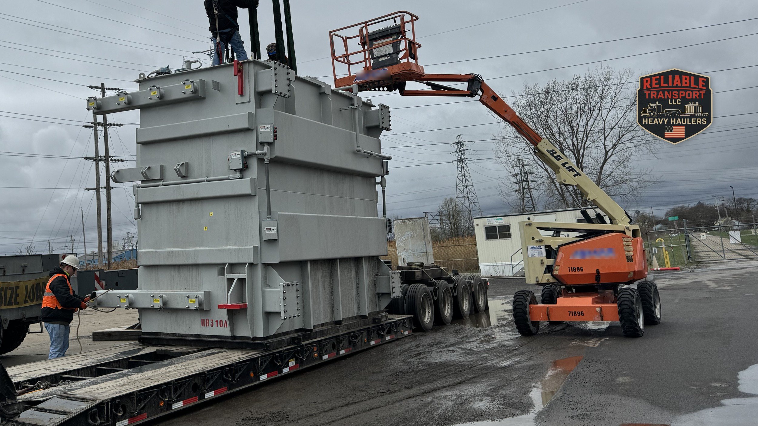 Crane lifting a gray electrical transformer onto a multi axle trailer under a cloudy sky