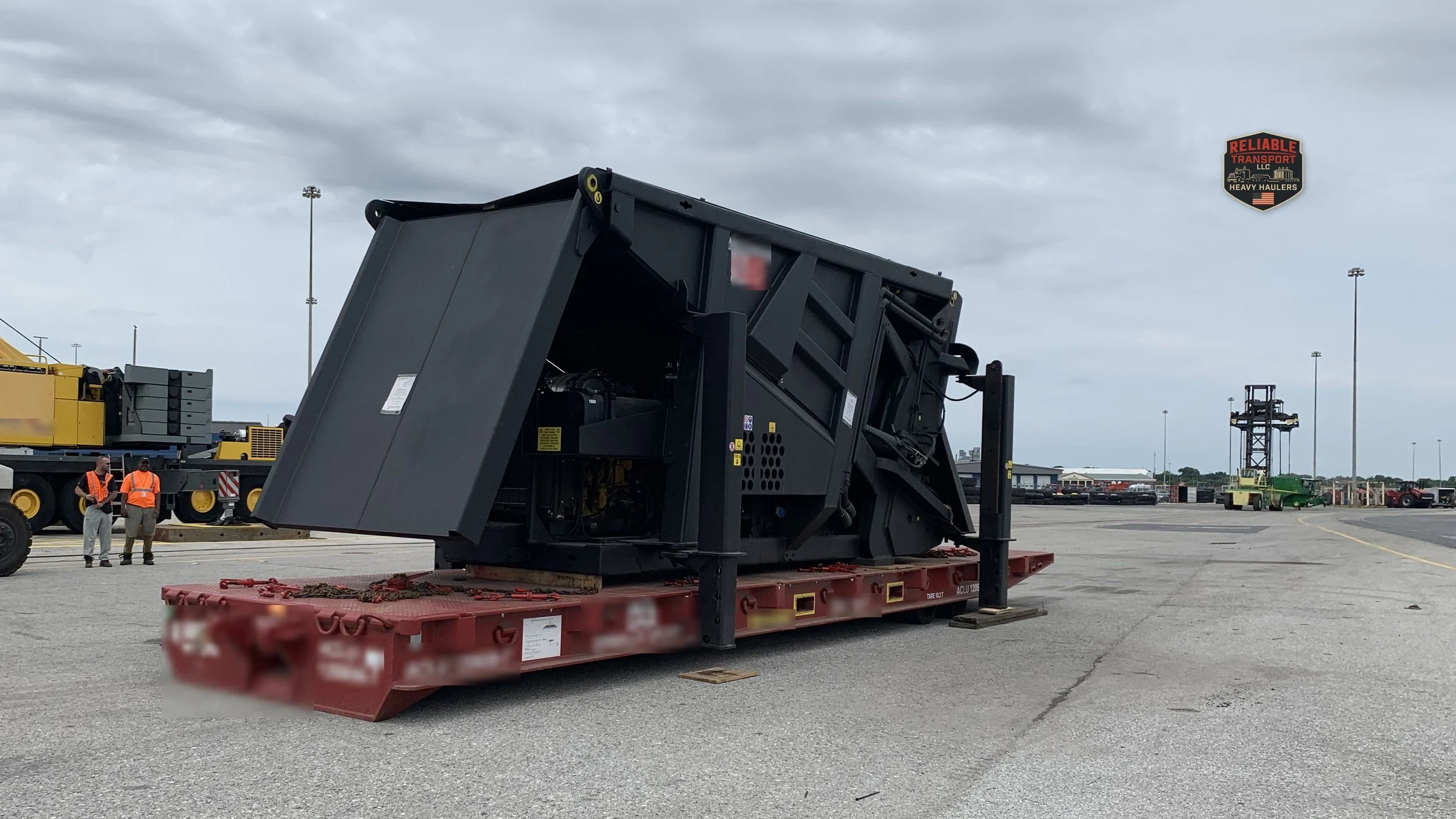 Large black steel machine on a red roll trailer in a wide paved yard under a cloudy sky