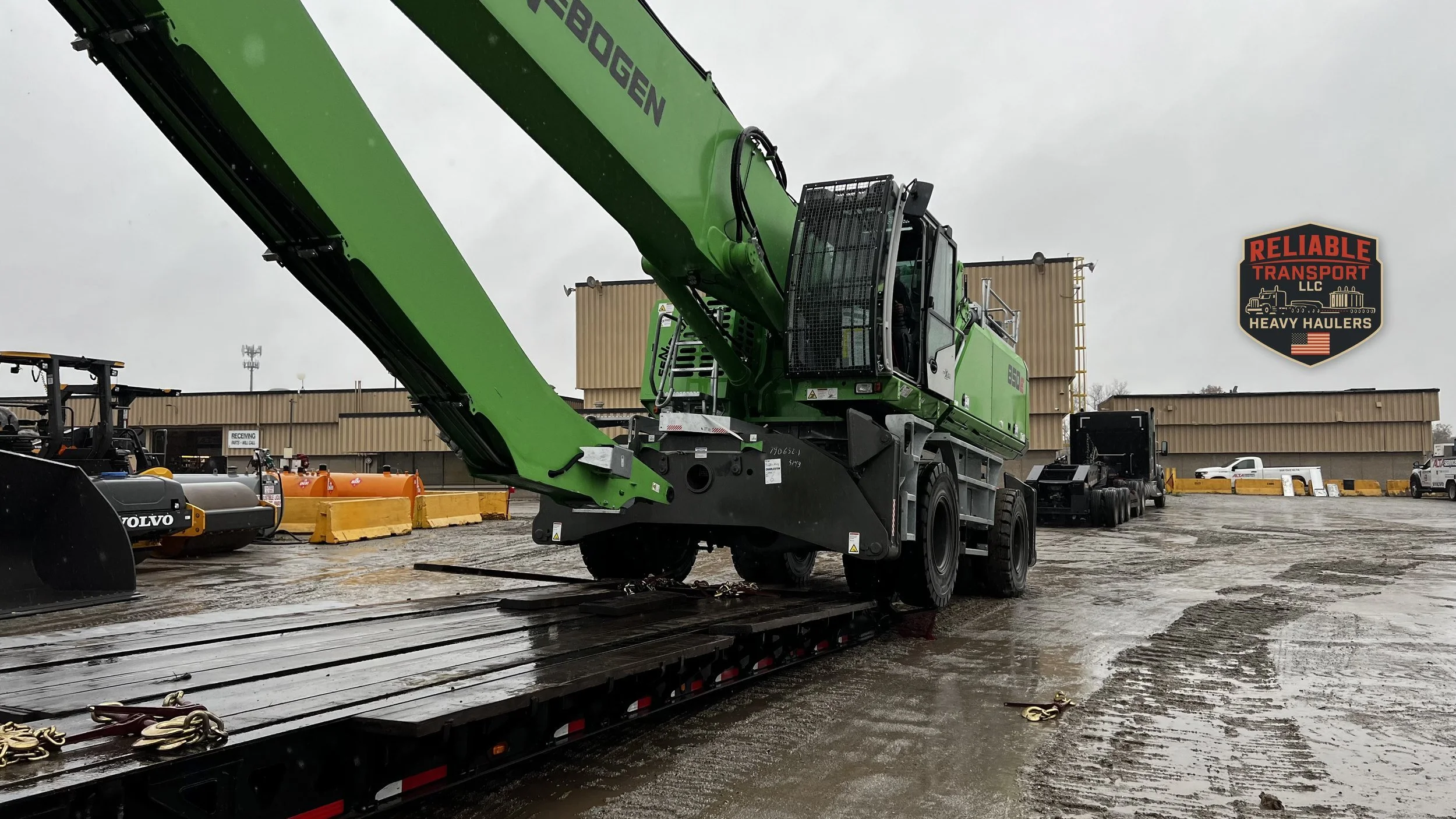 Green material handler loaded on a multi axle trailer in a wet industrial yard under a gray sky