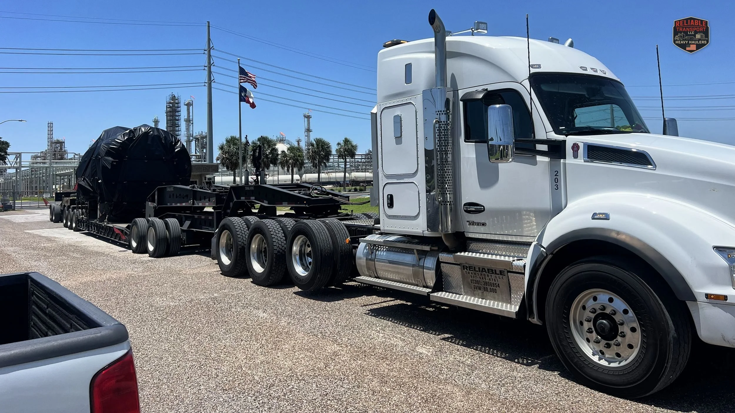 White heavy haul tractor pulling a tarped load on a multi axle trailer beside an industrial plant
