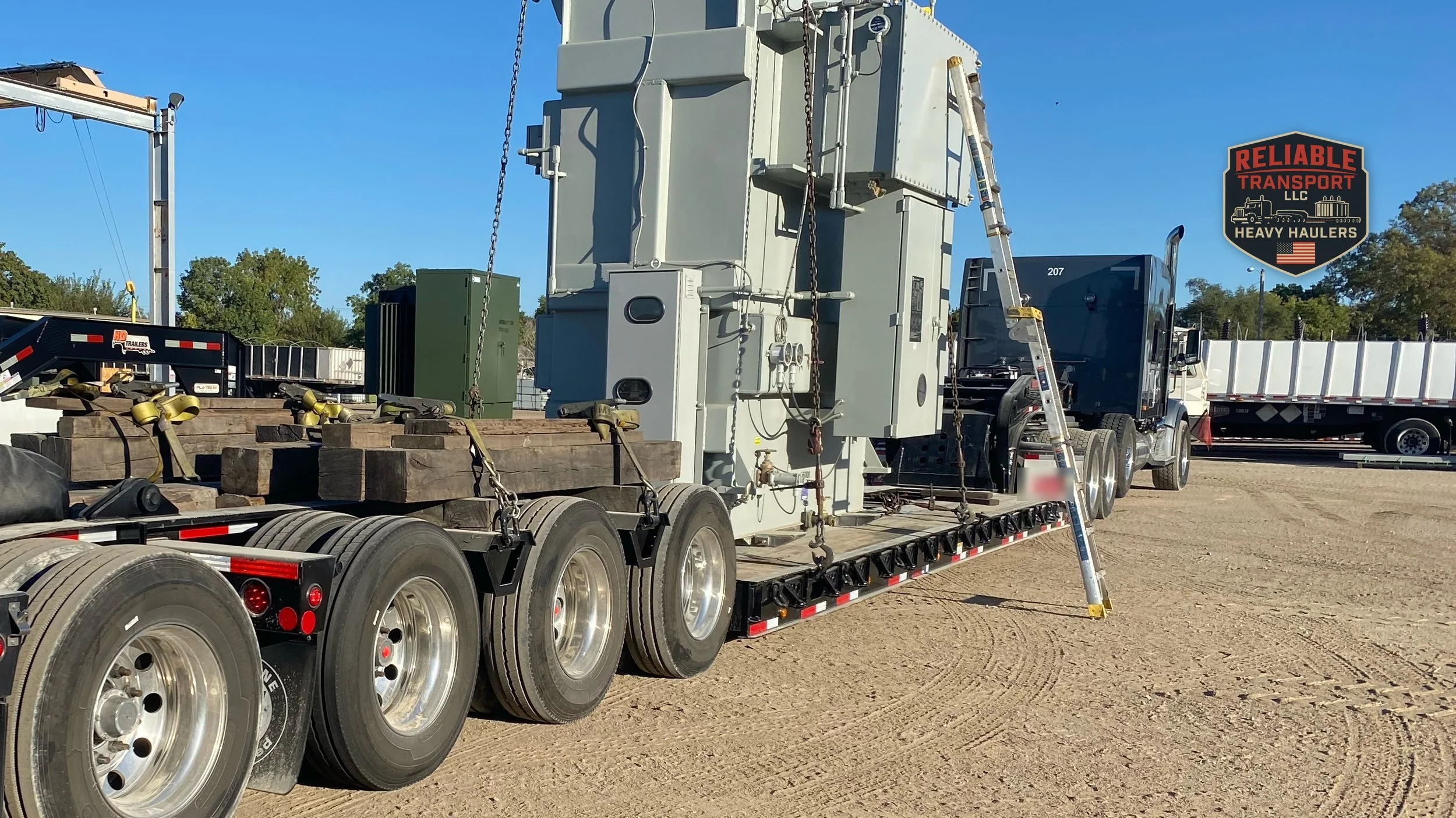 Gray electrical transformer chained on a multi axle trailer in a gravel yard under a blue sky