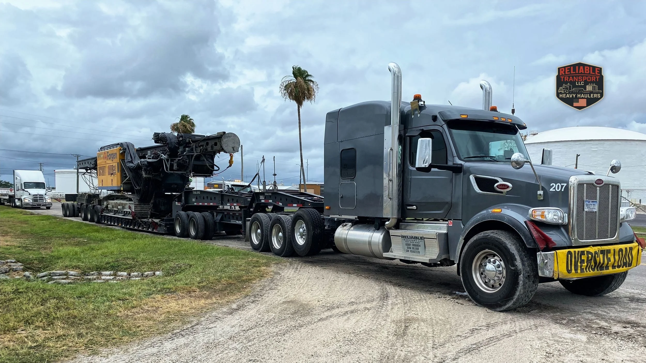 Gray heavy haul tractor pulling a tracked machine on a multi axle trailer under a cloudy sky