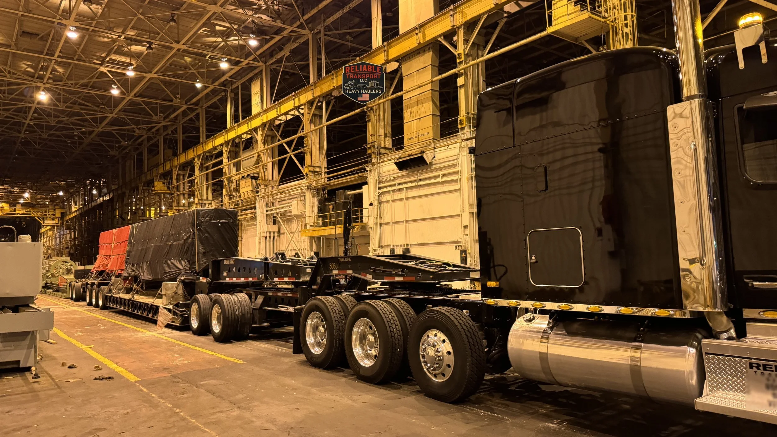 Black heavy haul tractor with tarped loads on a long multi axle trailer inside a large industrial building