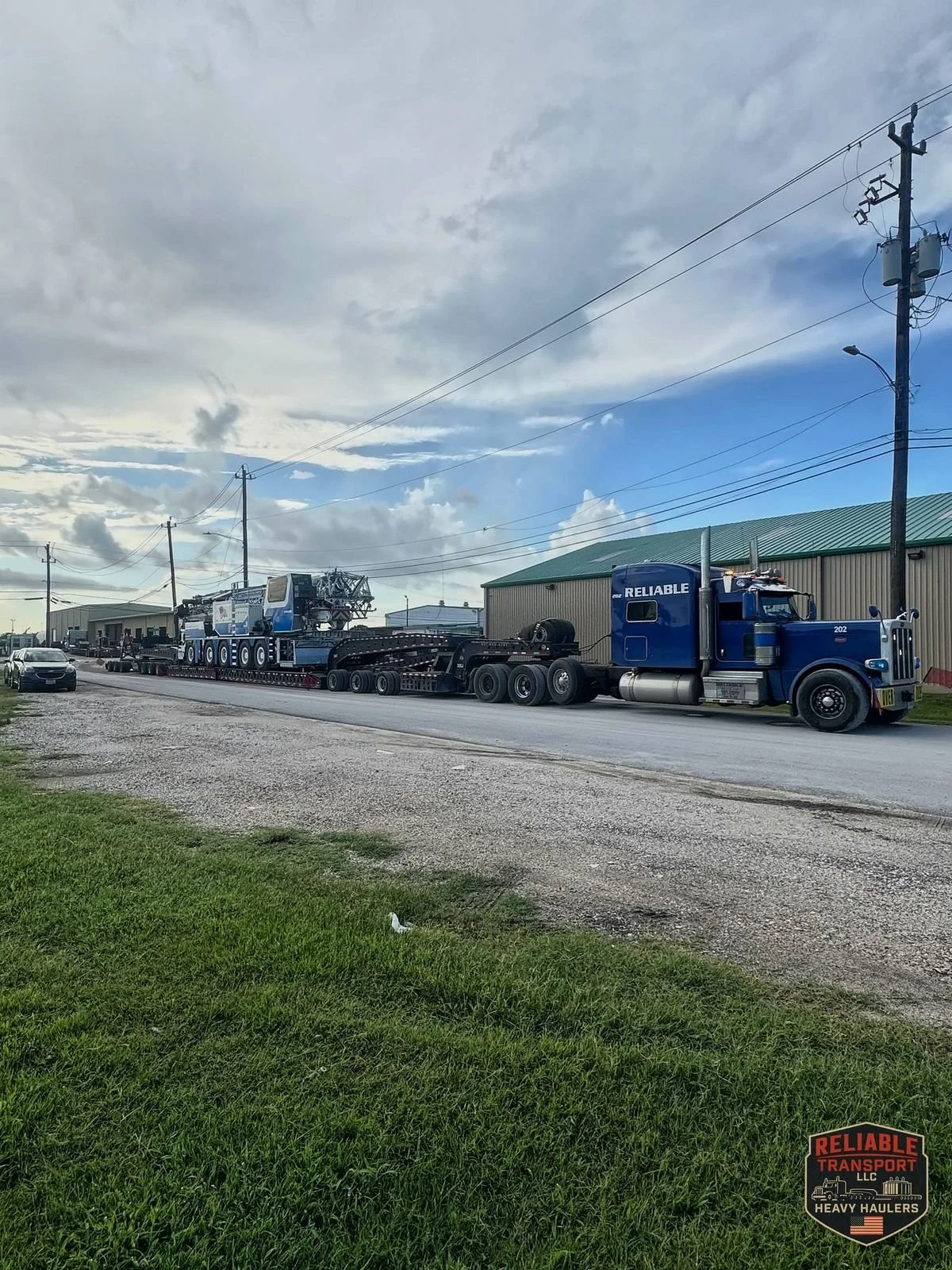 Blue Reliable truck pulling an all terrain crane on a long well trailer along a street beside a warehouse