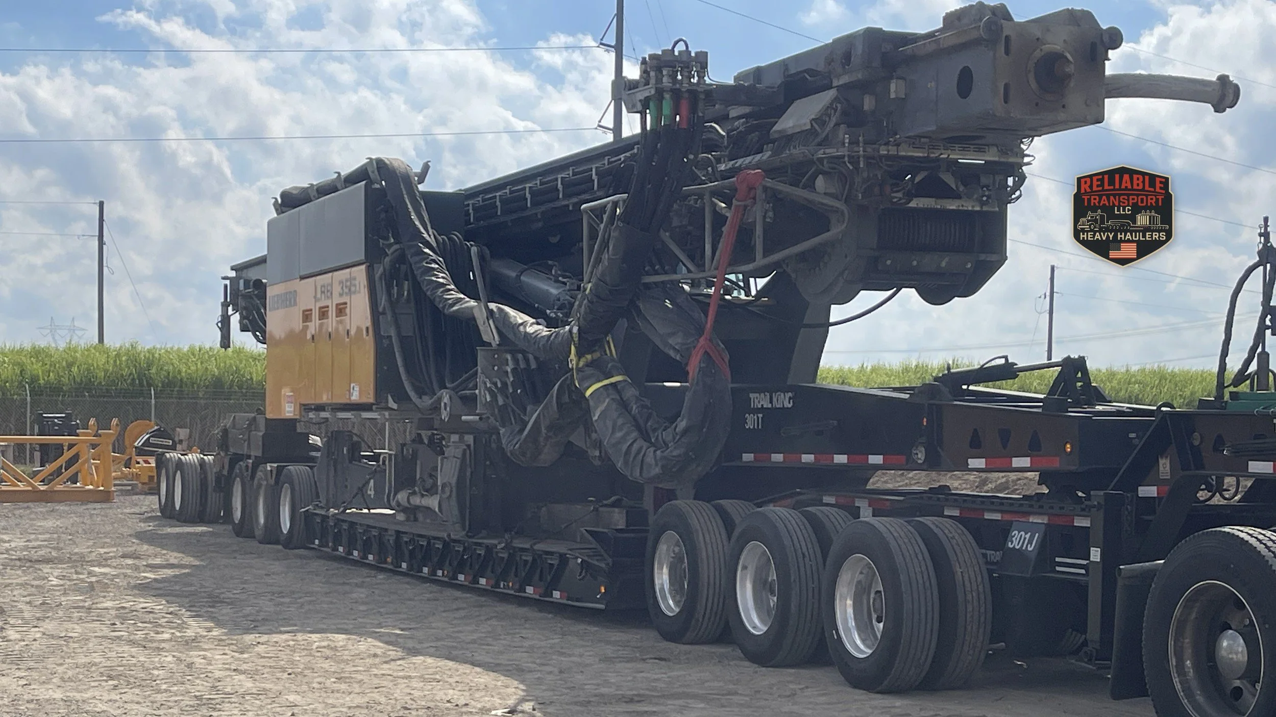 Yellow and black drilling rig chained to a multi axle trailer in a gravel yard under a cloudy sky