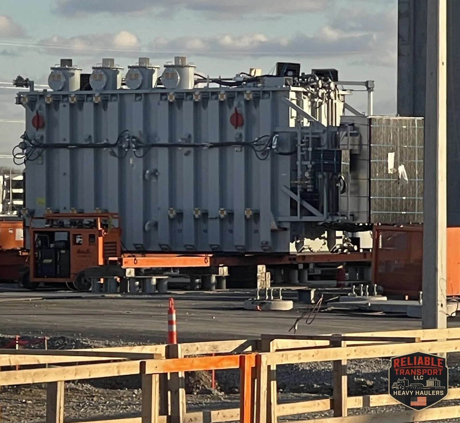 The 160,000 lb transformer on rigging equipment at the Obetz site in late afternoon light, behind a wooden safety fence