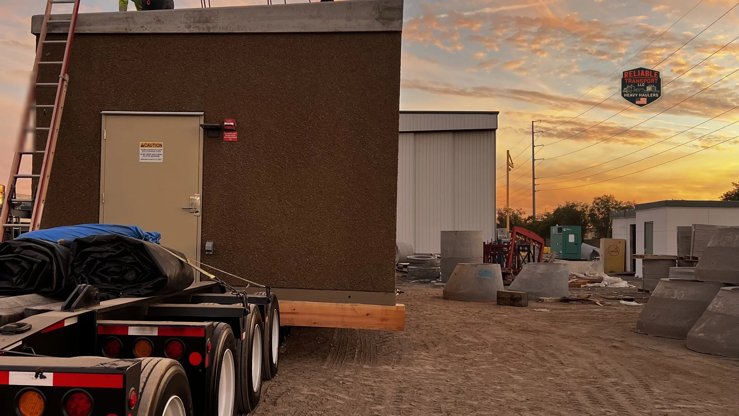 Brown modular building on a multi axle trailer in an industrial yard under a sunset sky