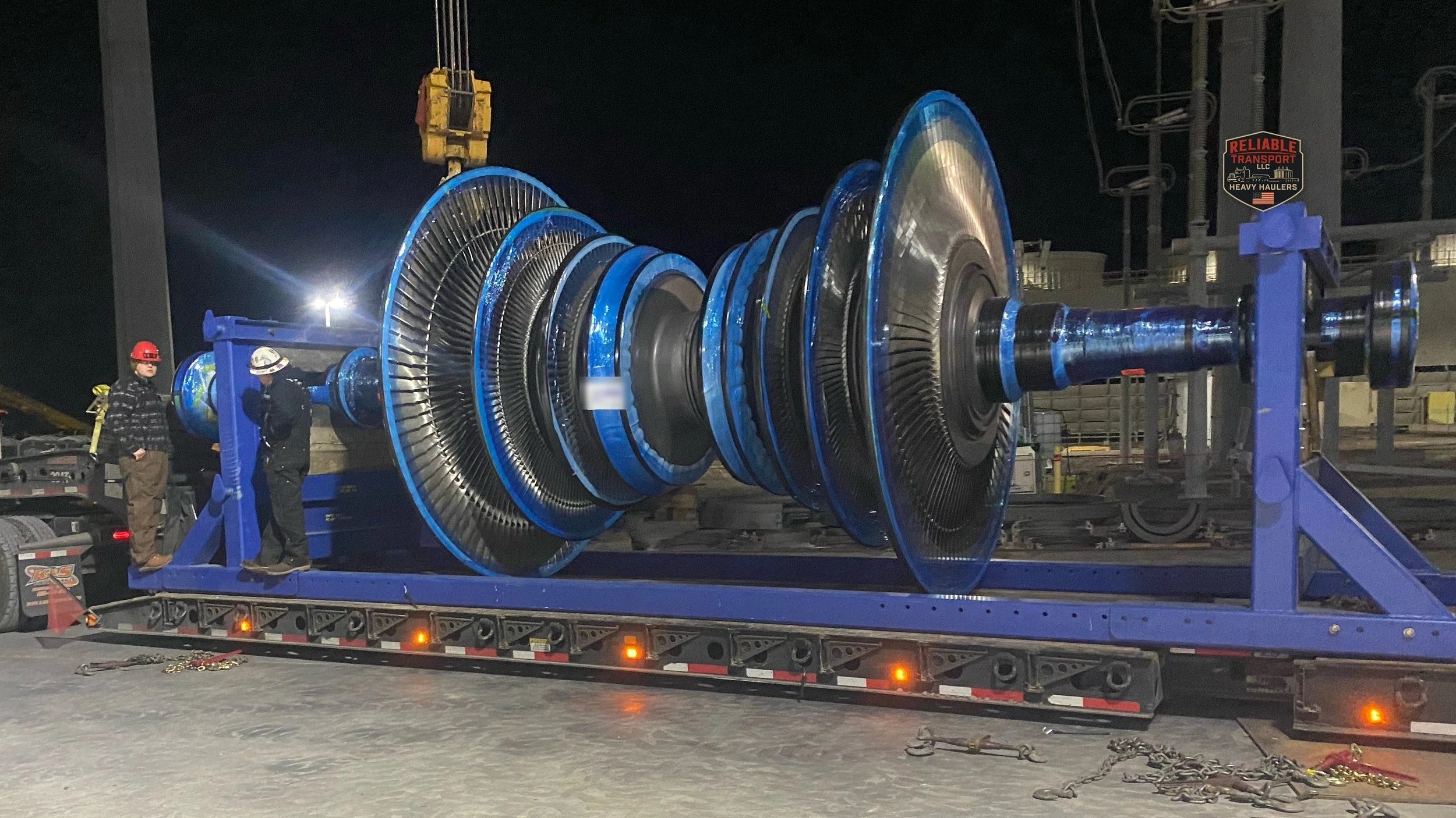 Blue turbine rotor on a steel cradle atop a multi axle trailer at night