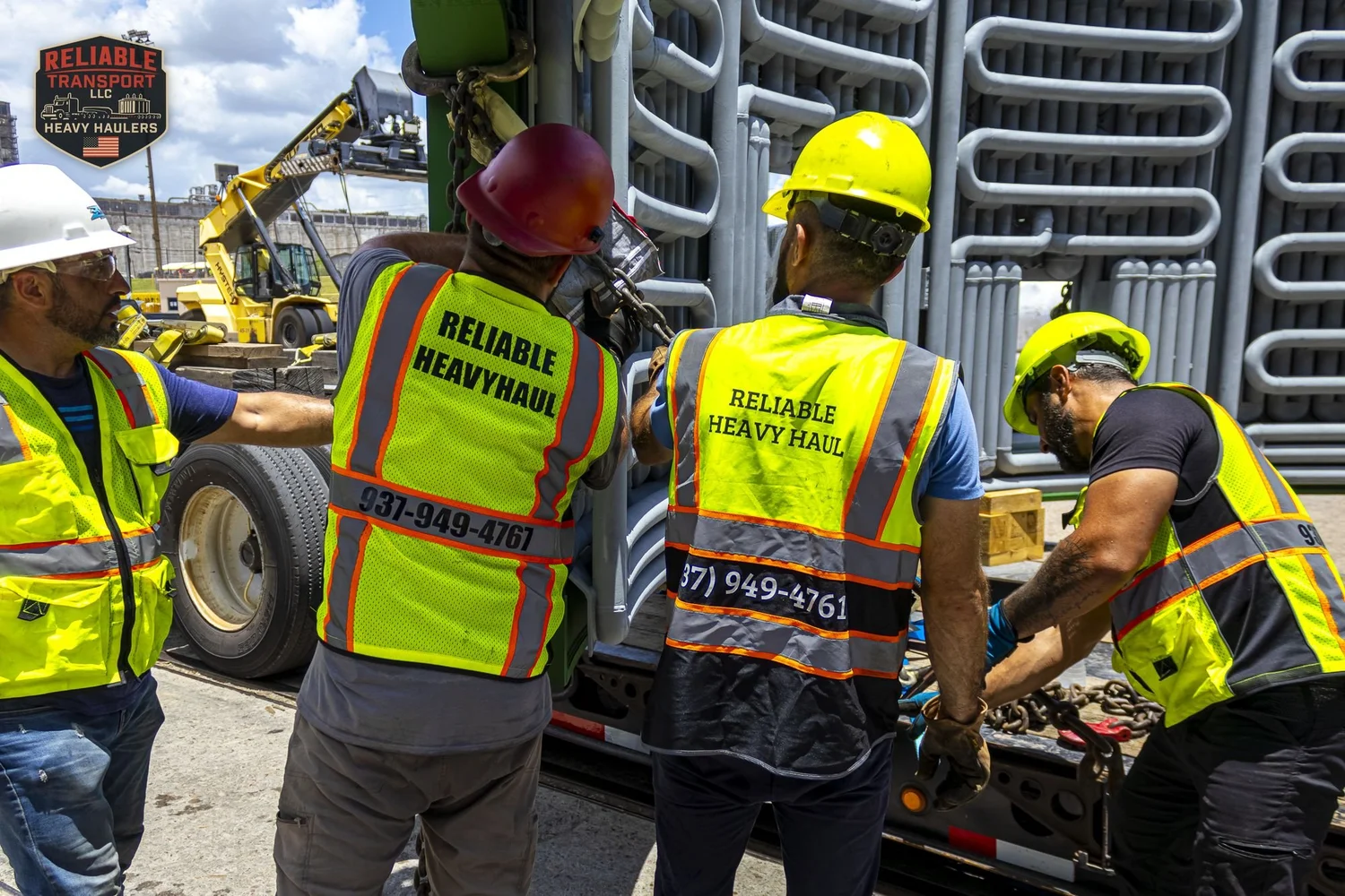Crew in hard hats and high visibility vests rigging chains on a wide load at the trailer