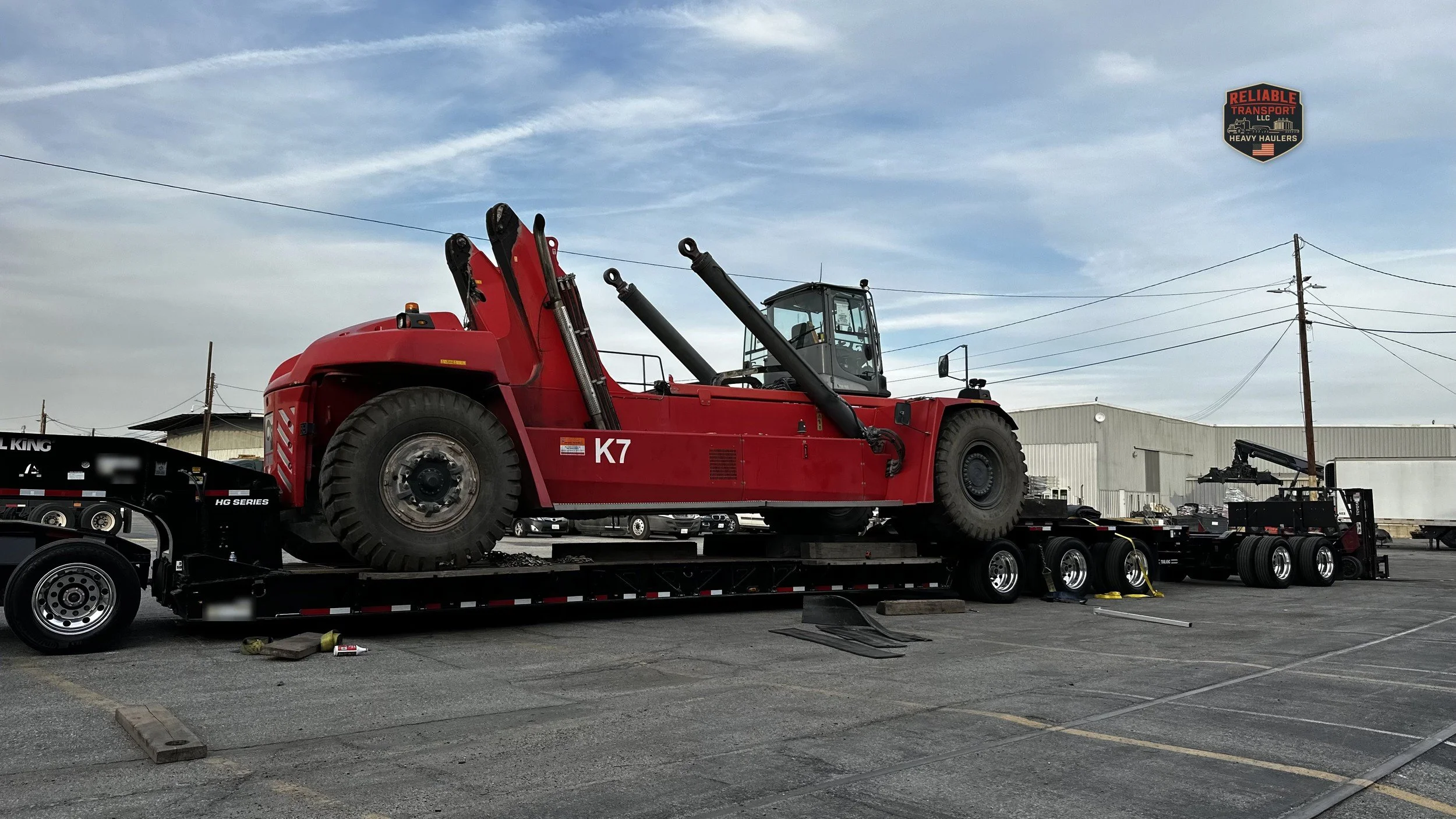 Red container handler loaded on a multi axle trailer in a paved lot under a blue sky