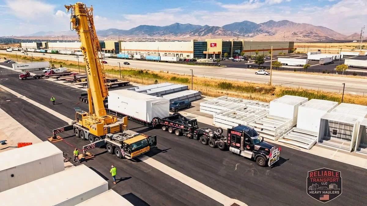 Drone view of a yellow mobile crane set up beside a black Reliable Transport truck with a white module on its trailer, mountains behind